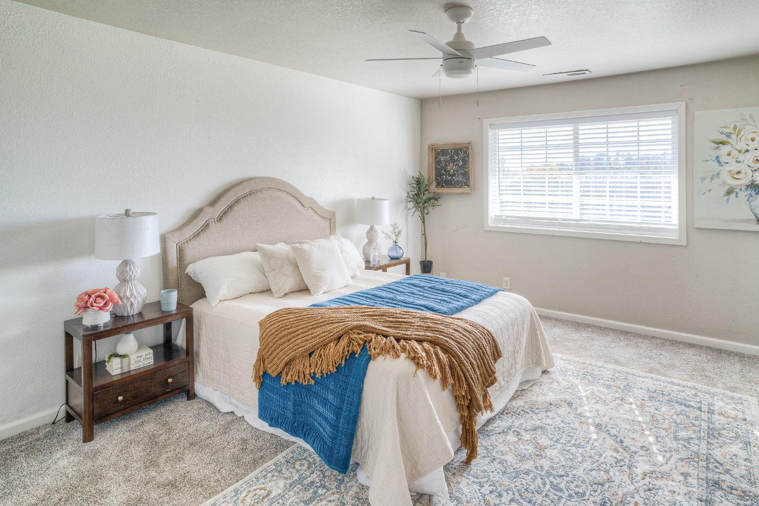Furnished bedroom with a blue-and-tan layered bed, upholstered headboard, and large window with white blinds.