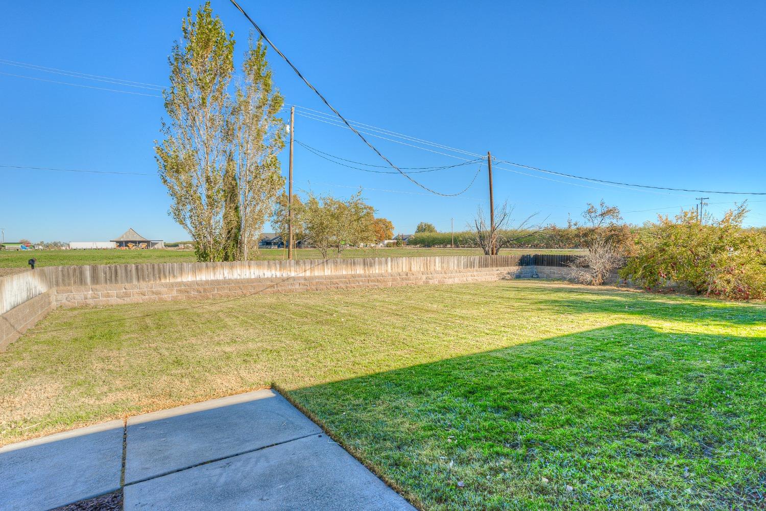 Backyard lawn bordered by a low retaining wall and fence, with a concrete patio and open fields beyond.
