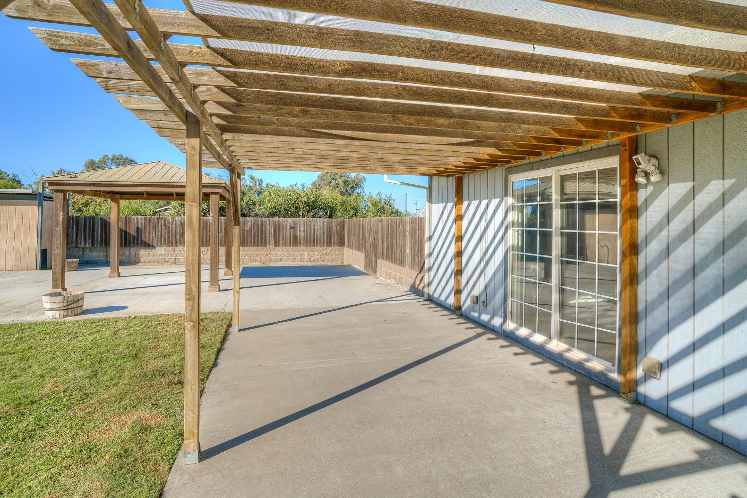 Fenced backyard patio with a broad wooden pergola, concrete paving, sliding glass doors, and a small lawn.