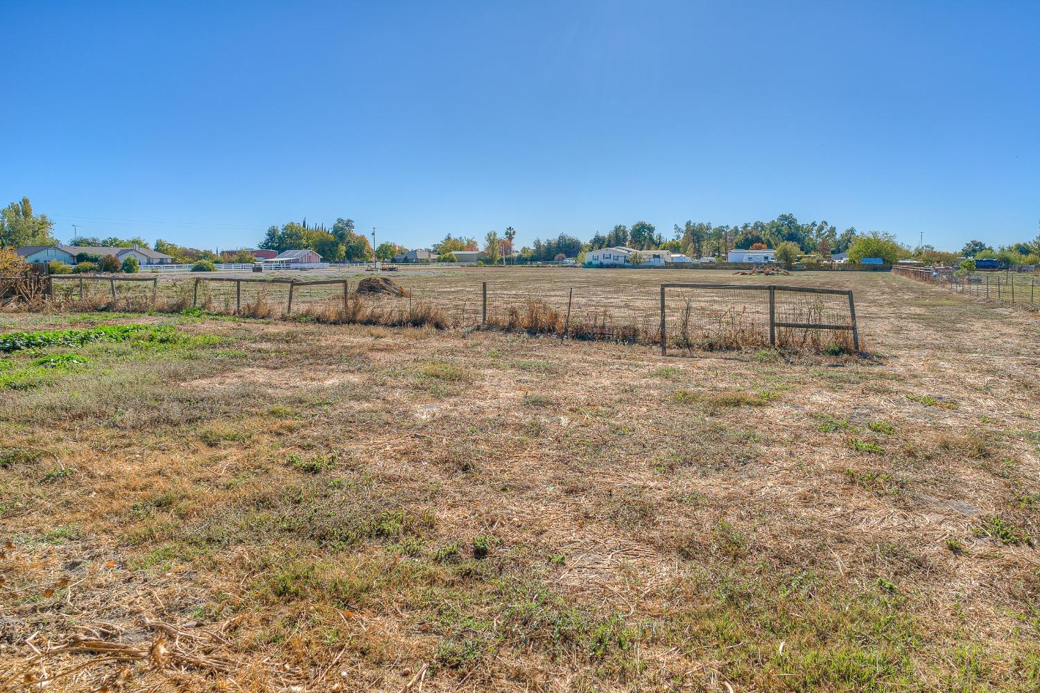 A broad, dry vacant lot enclosed by fencing, with homes and trees along the far edge.