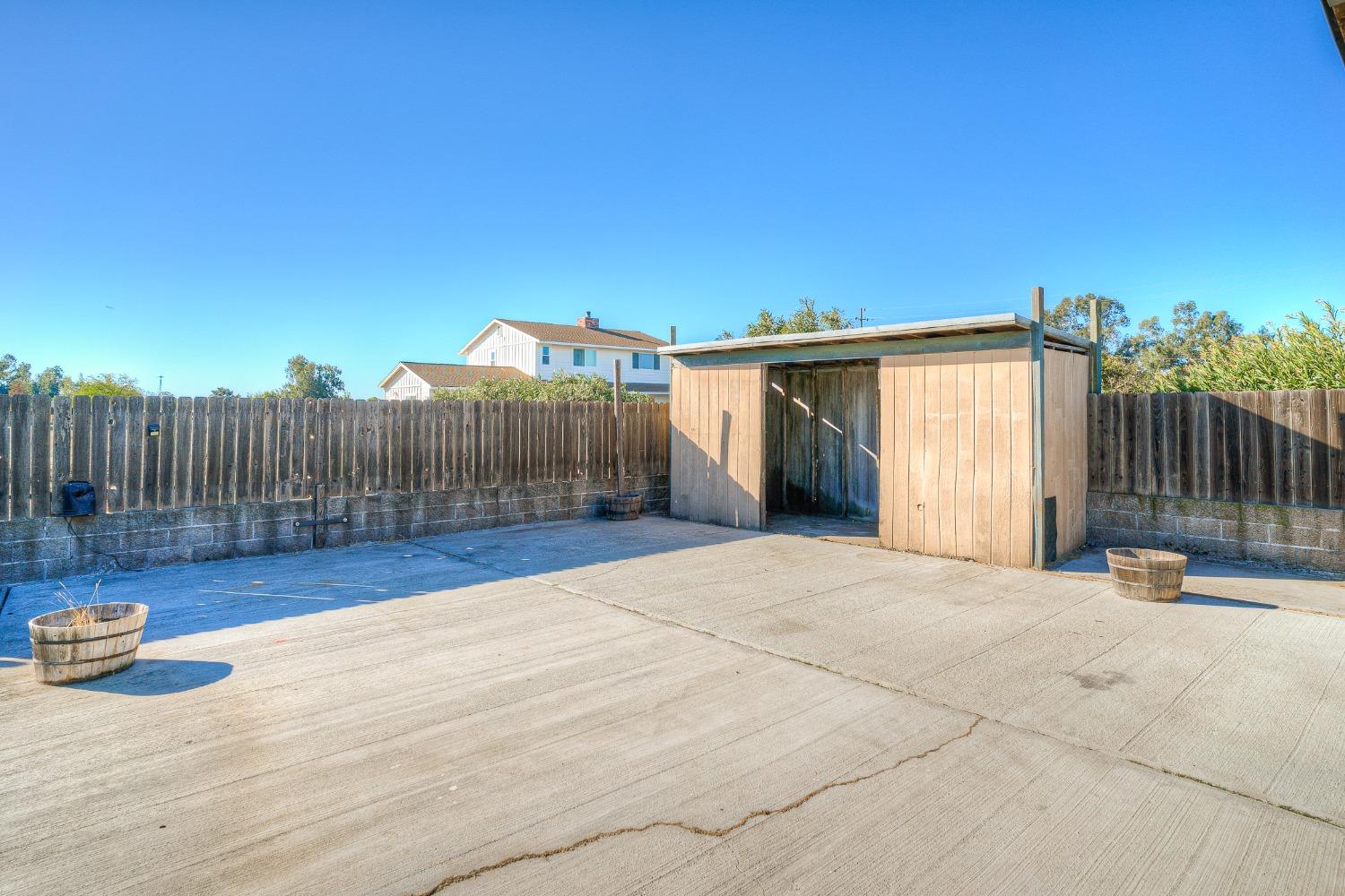 Concrete backyard patio with a wooden shed, tall fence, and barrel planters.