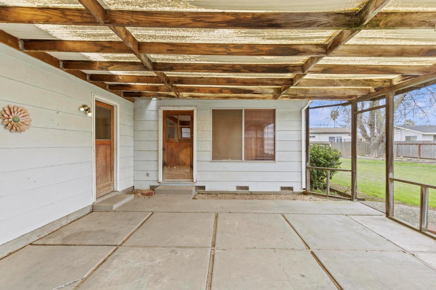 Covered concrete patio with exposed wooden beams, white siding, and a view of the fenced lawn.