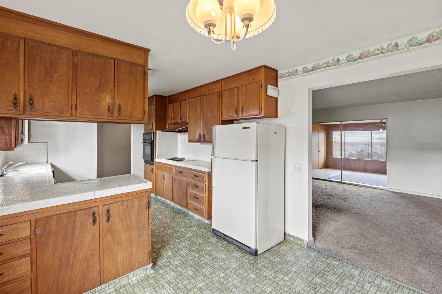 Kitchen with wood cabinets, white refrigerator, patterned green floor, and an adjoining carpeted room.