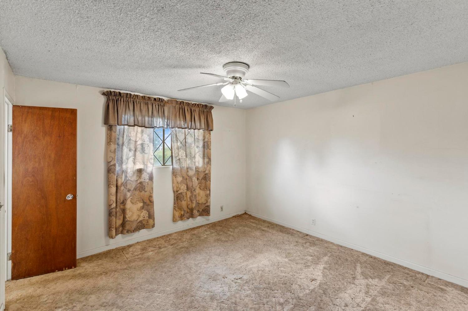 Empty bedroom with beige carpet, a ceiling fan, and patterned curtains over the window.