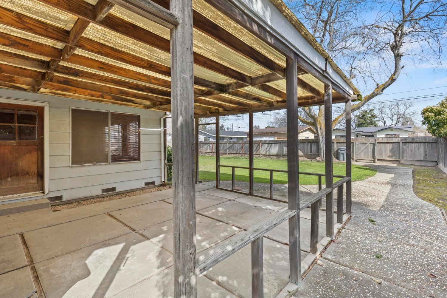 Covered backyard patio with a wooden pergola, concrete paving, and a path beside the fenced lawn.