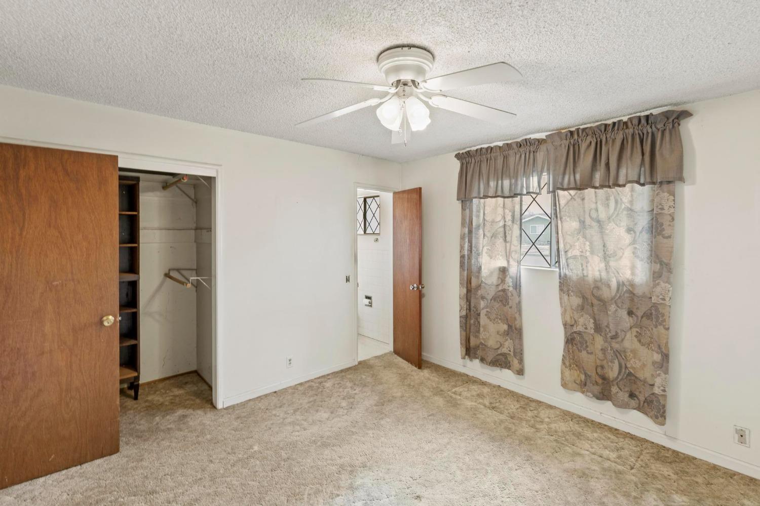 Empty bedroom with beige carpet, ceiling fan, curtained window, and open closet.