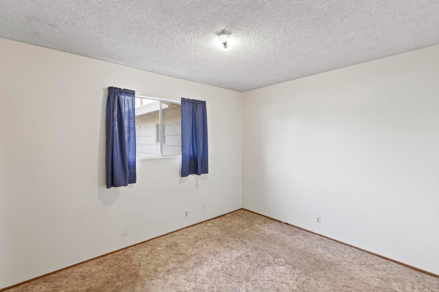 Empty carpeted room with white walls and a window framed by blue curtains.