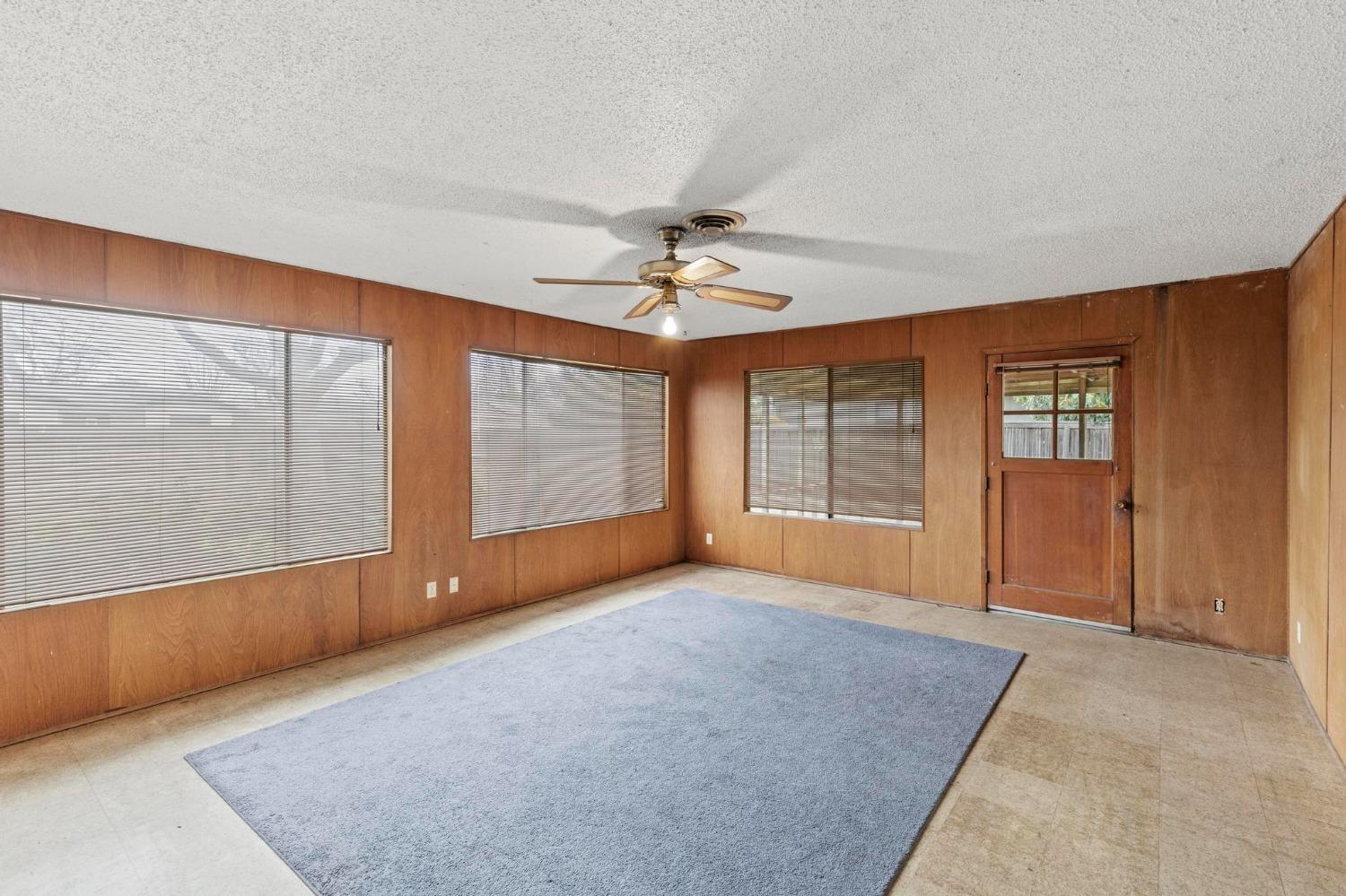 Empty living room with wood-paneled walls, window blinds, a ceiling fan, and a large gray rug.