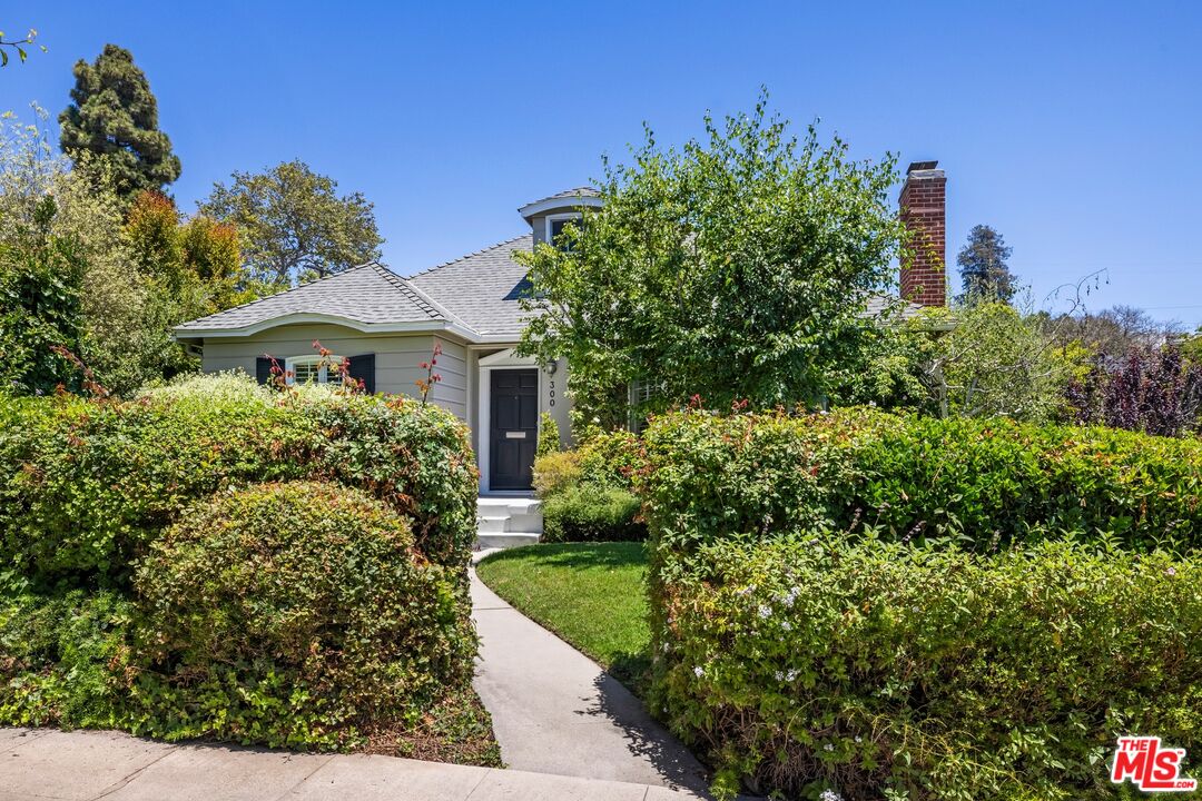 Gray house with a dark front door and brick chimney, approached by a curved walkway through landscaped hedges.