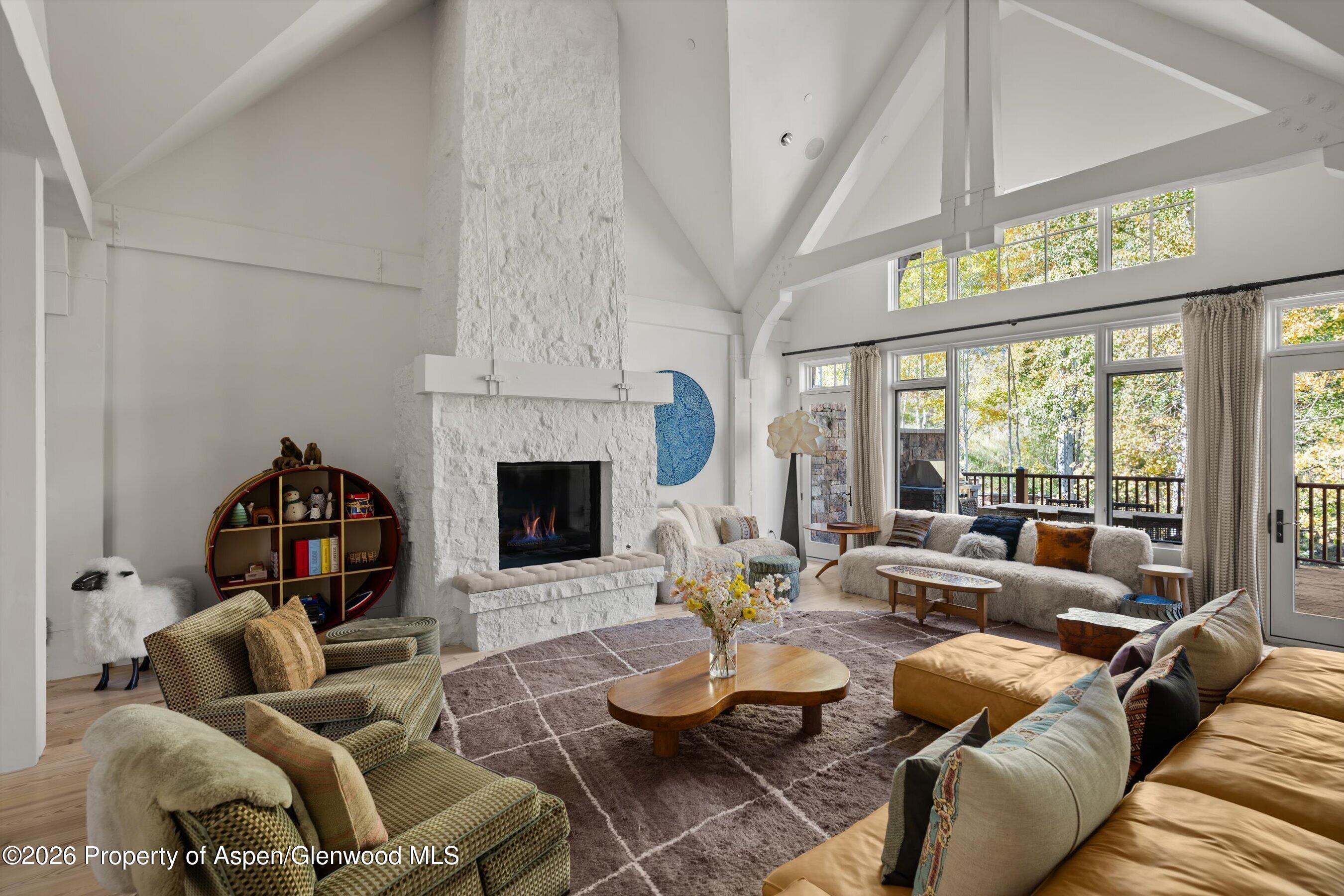 Living room with vaulted beamed ceiling, white stone fireplace, and tall windows overlooking autumn trees.