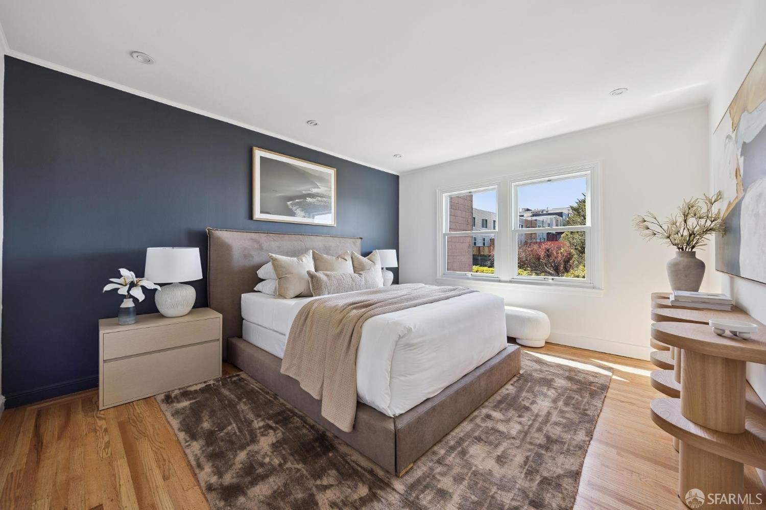 Staged bedroom with a navy accent wall, white bedding, hardwood floors, and bright windows.