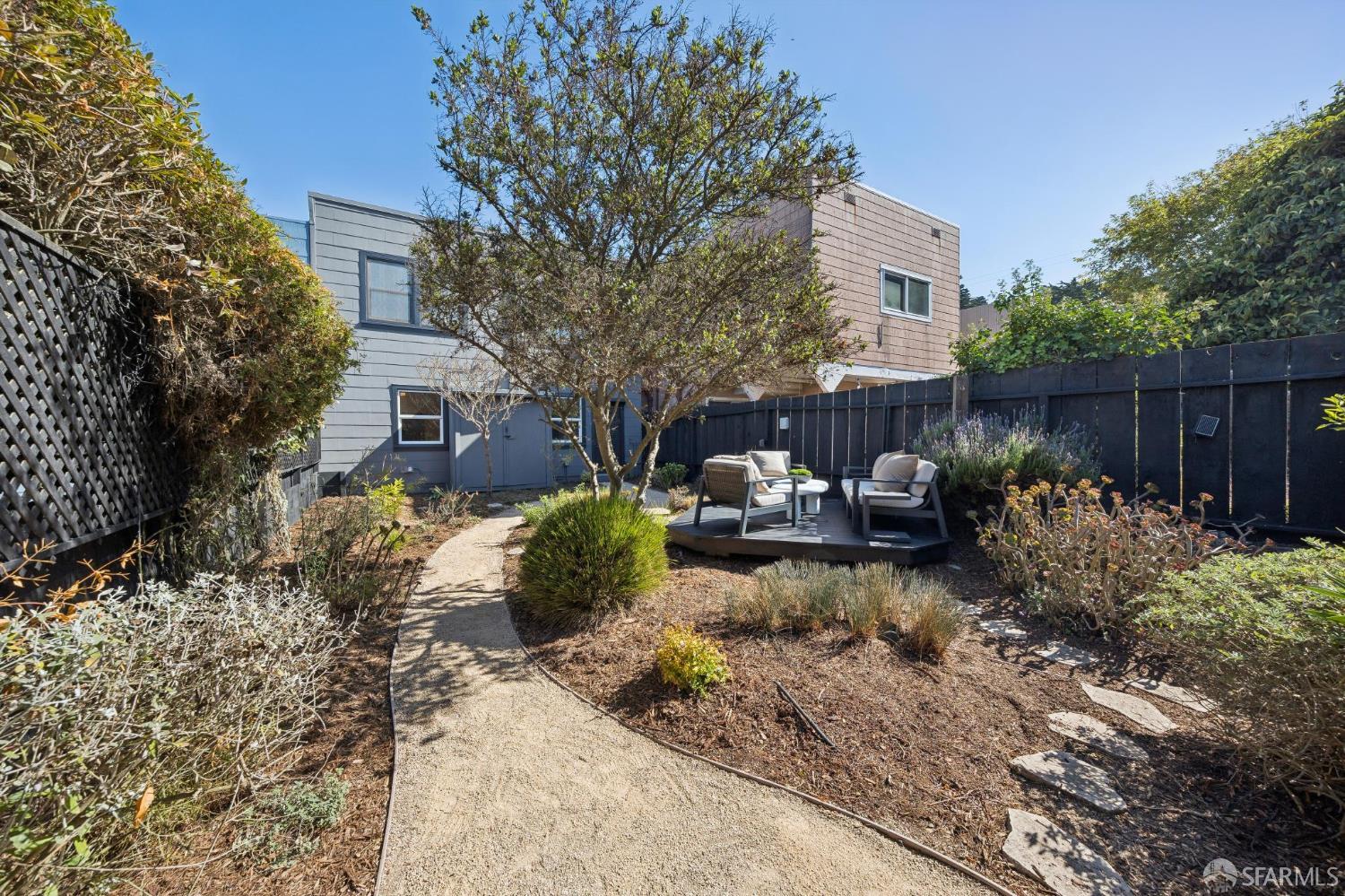 Backyard garden path leads to a raised patio with outdoor seating beside a modern two-story home.