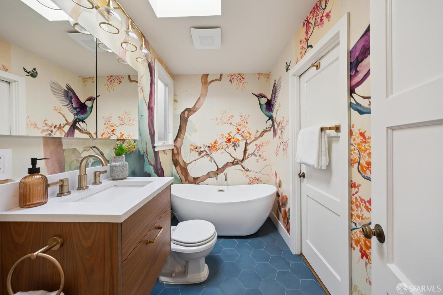Bathroom with a freestanding tub, blue hexagonal tile, wood vanity, and colorful bird mural wallpaper.