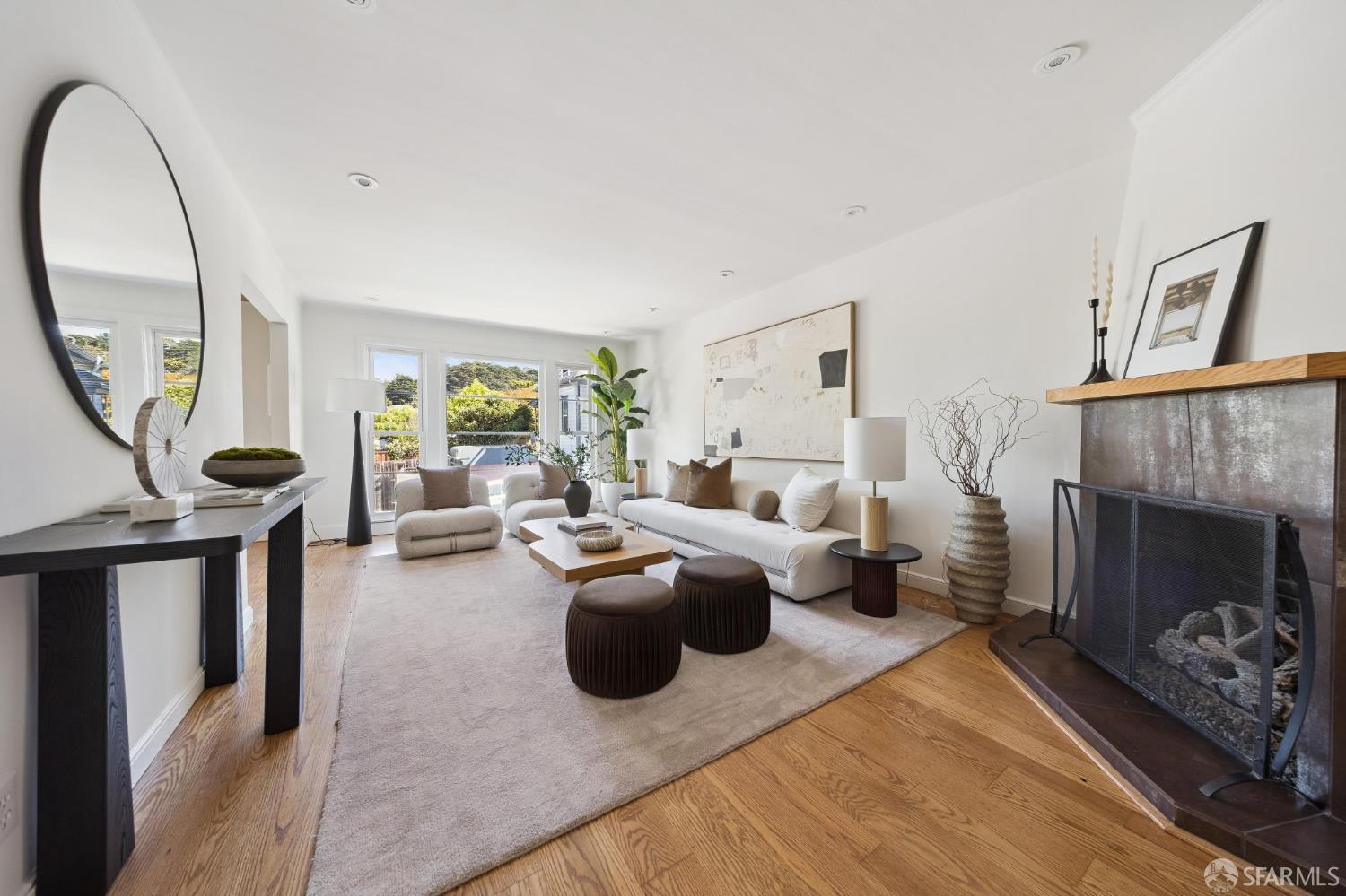 Staged living room with a white sofa, hardwood floors, large windows, and a fireplace.