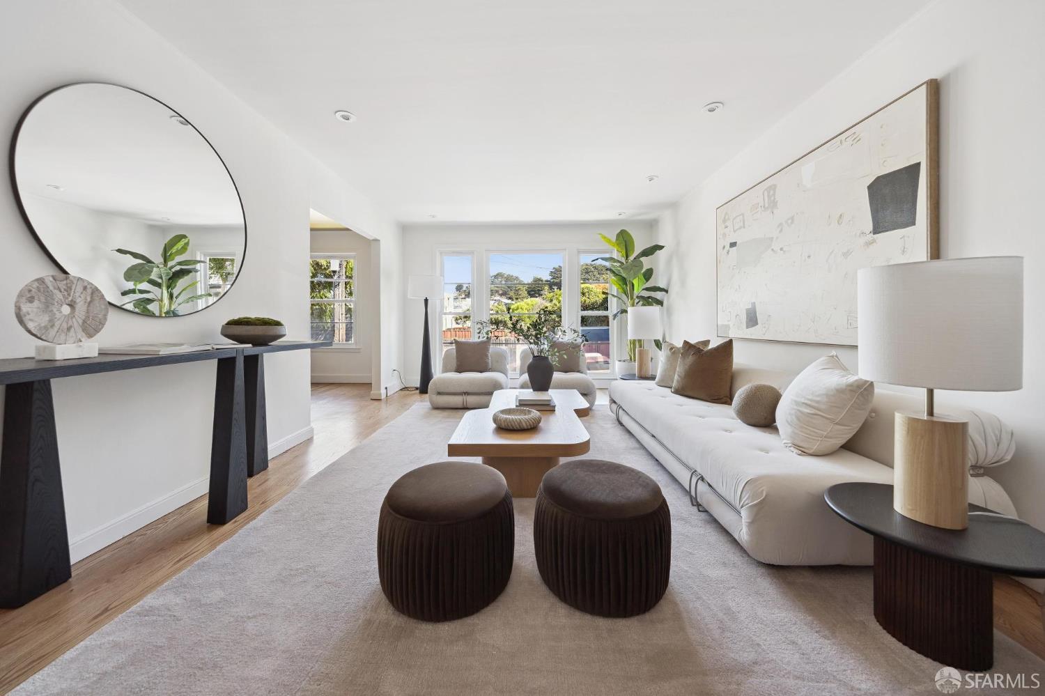 Staged living room with a long white sofa, round ottomans, and wide windows overlooking greenery.