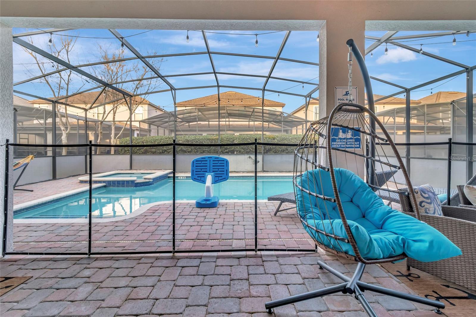 Screened backyard pool with a basketball hoop and turquoise-cushioned hanging chair on the paver deck.