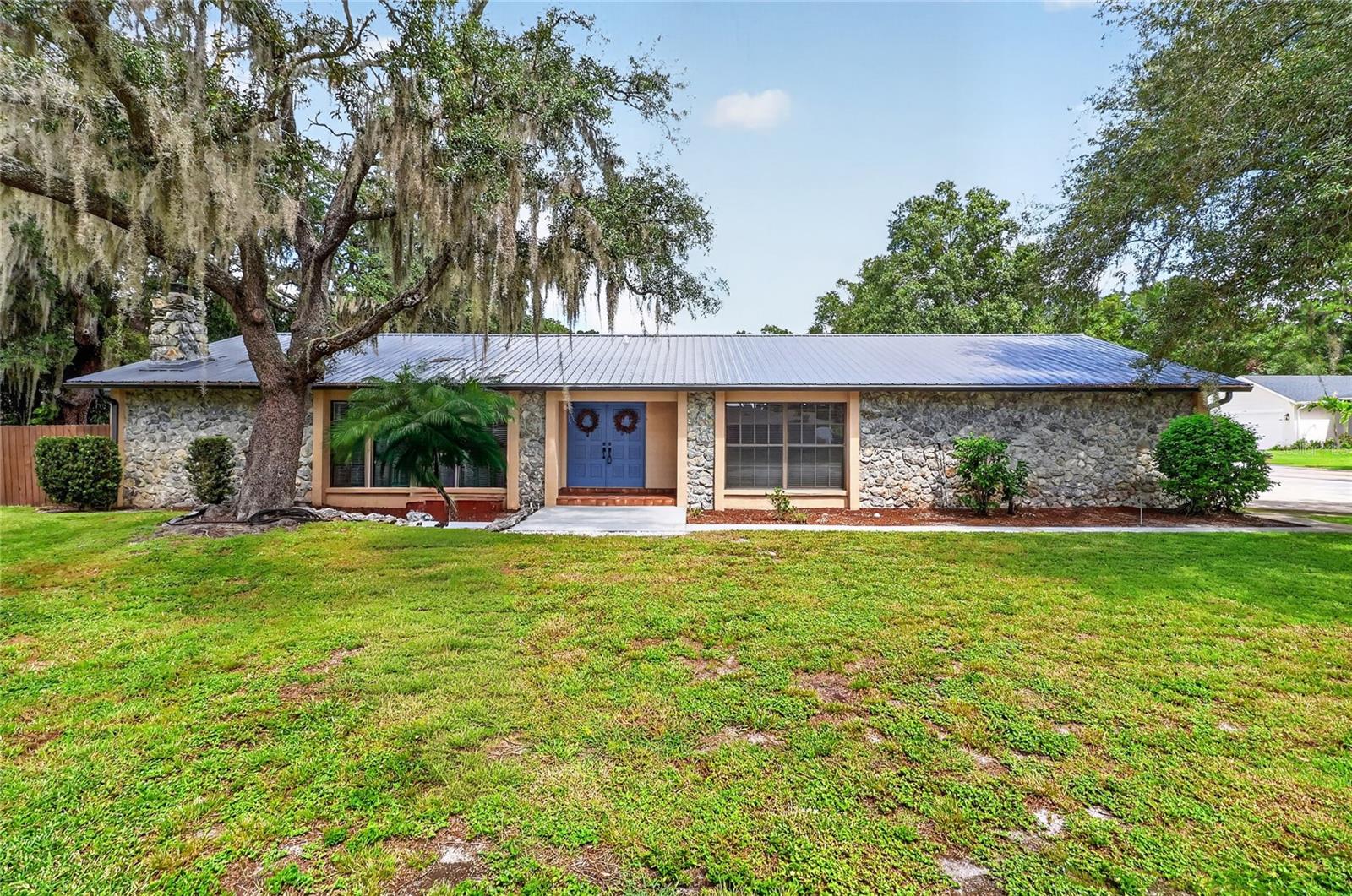 Single-story stone-front house with a gray metal roof, blue double doors, and a moss-draped oak tree.