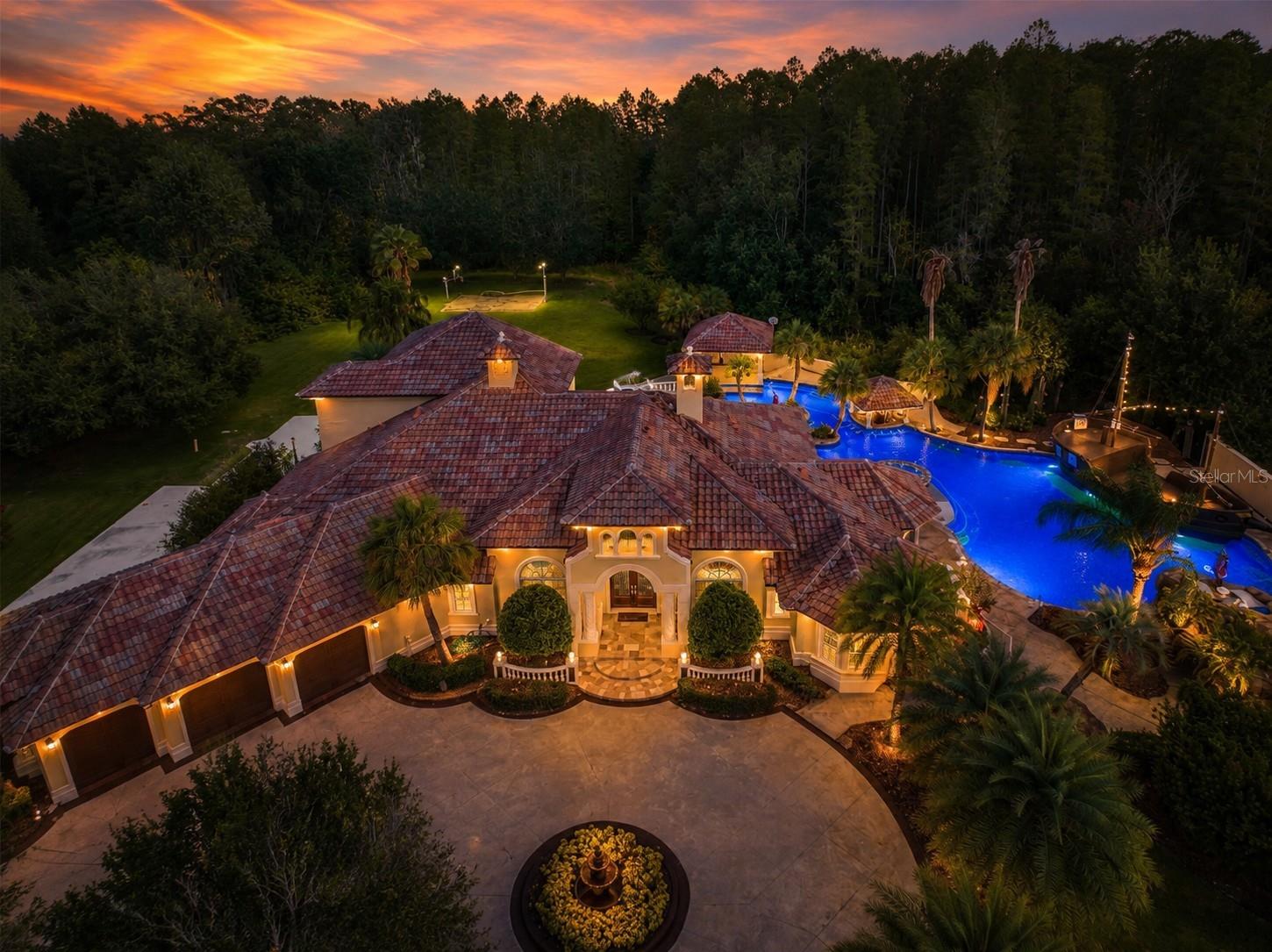 Aerial view of a large tiled-roof home with an illuminated pool and landscaped courtyard at sunset.