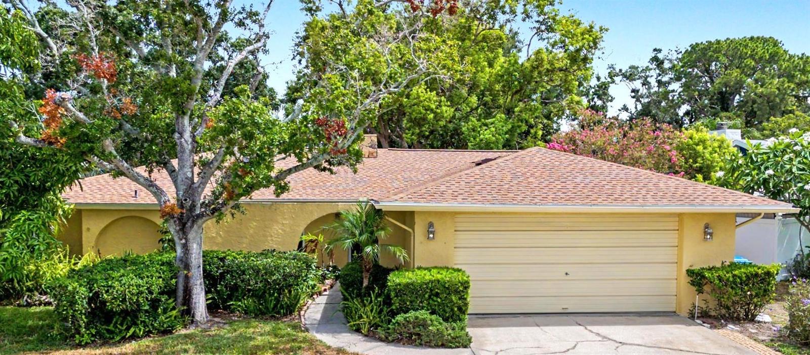 Yellow single-story home with a two-car garage, curved walkway, hedges, and mature trees.