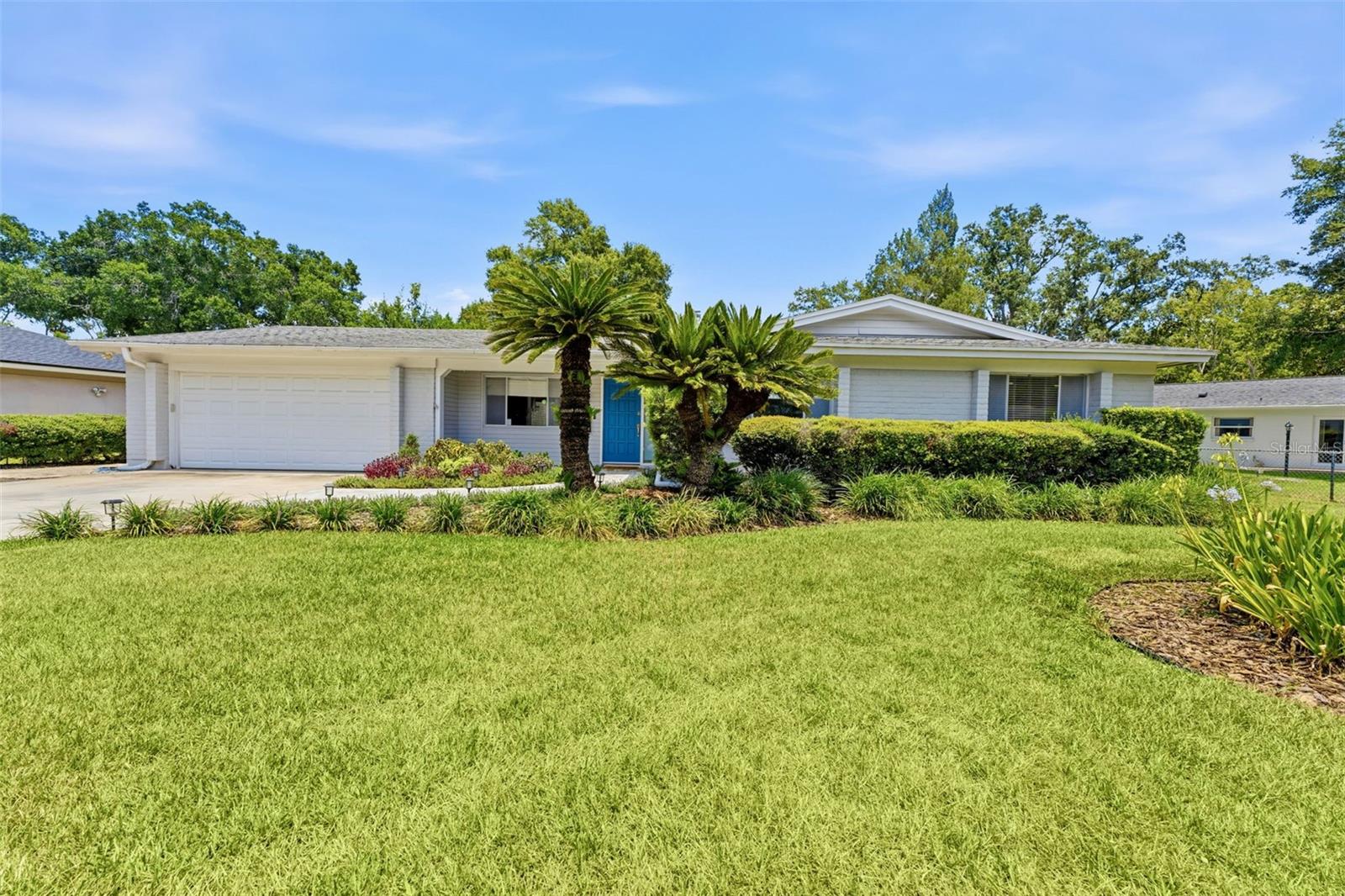 Single-story white home with a blue front door, two-car garage, palm trees, and a broad lawn.