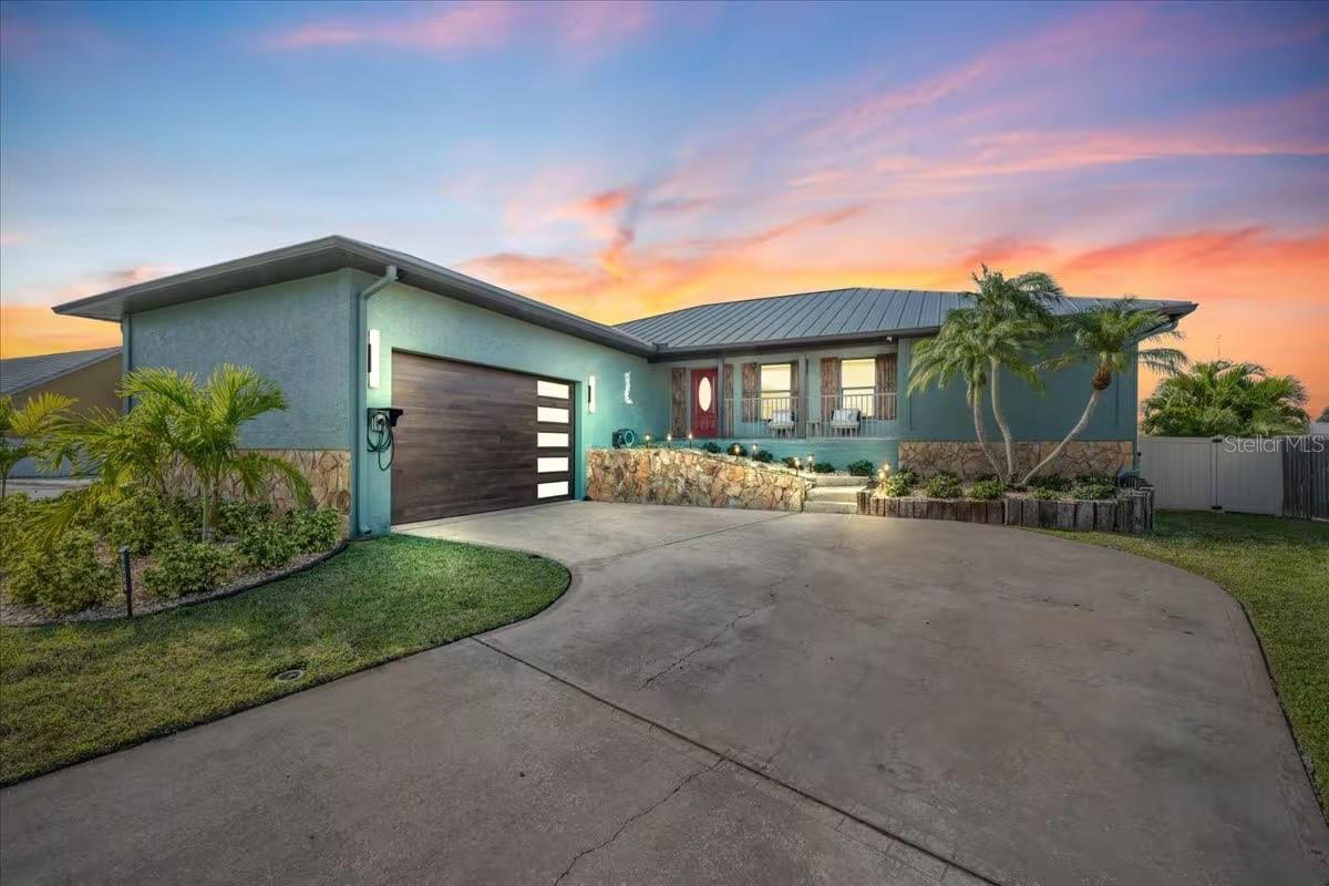 Modern single-story home with a wide driveway, stone landscaping, palm trees, and a pink-orange sunset.