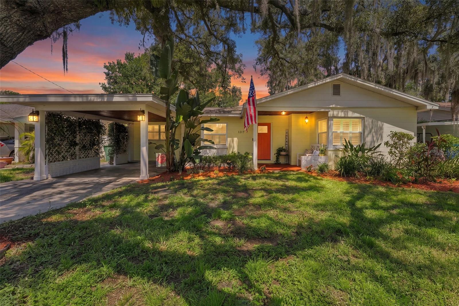 Single-story home with a red front door and carport beneath oak trees at sunset.