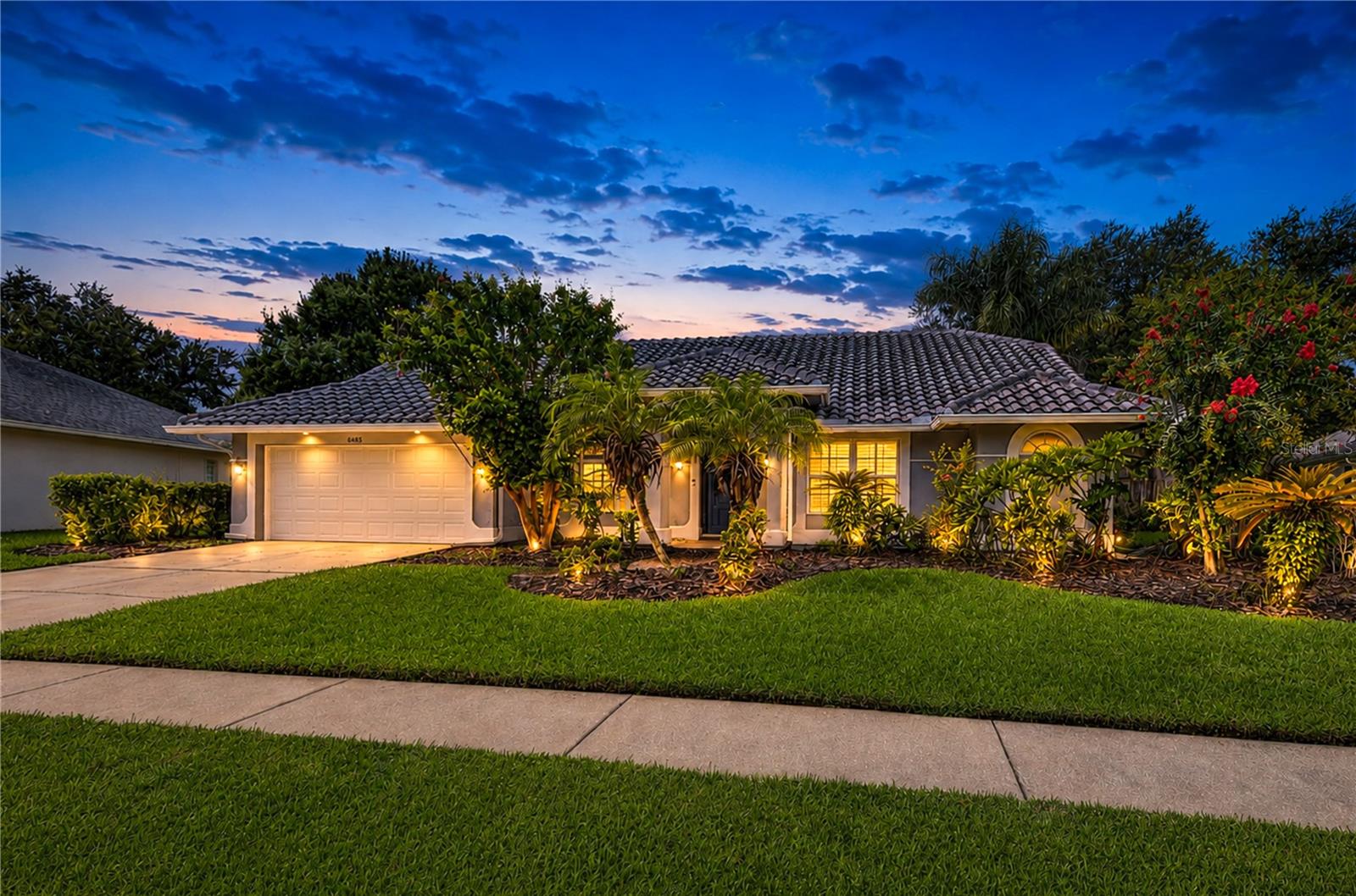 Single-story home with a tiled roof, two-car garage, and tropical landscaping at dusk.