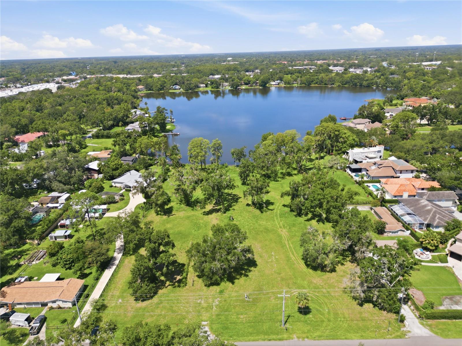 Vacant grassy lot beside a lake, surrounded by trees and homes.