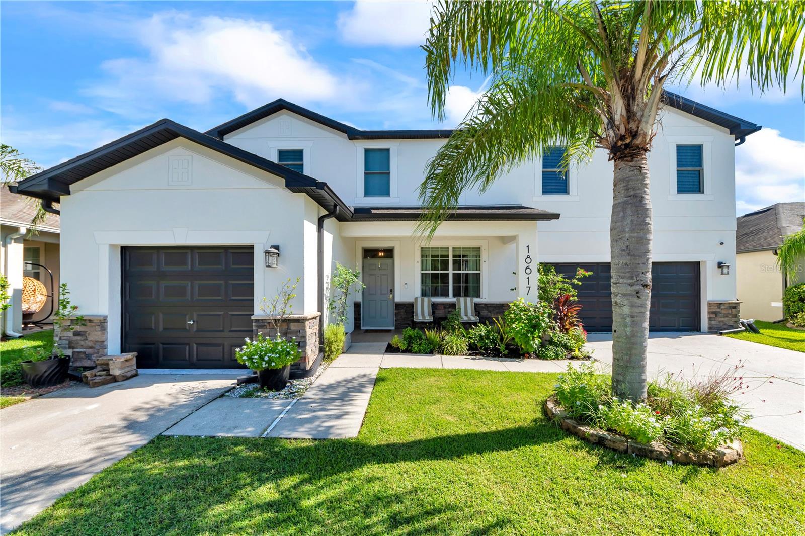 Two-story white house with dark double garages, landscaped lawn, and a palm tree.