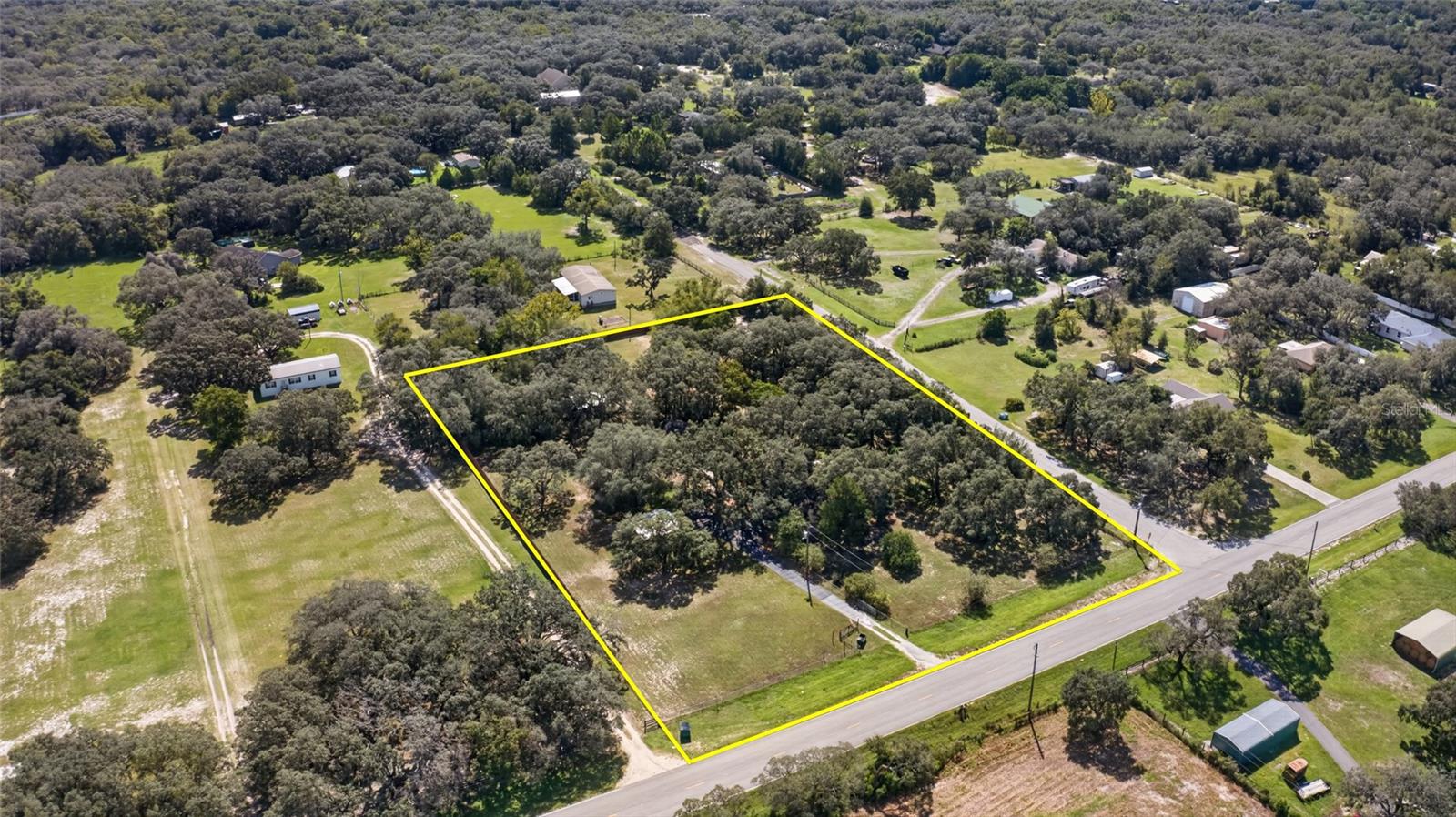Aerial view of a wooded rural lot outlined in yellow, with open grass and road frontage.