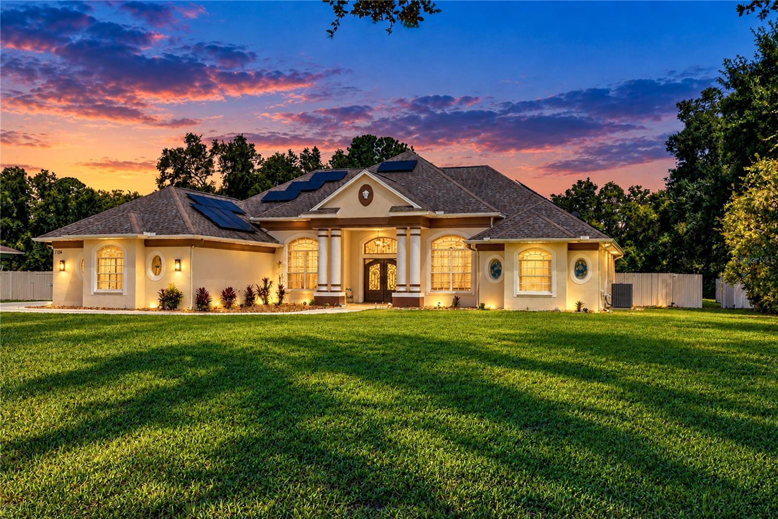 Single-story cream house with solar panels and illuminated windows beneath a colorful dusk sky.