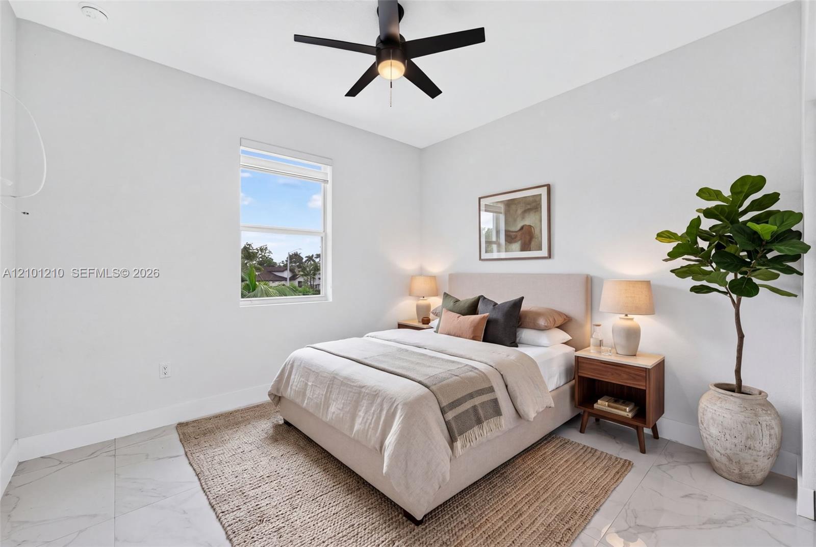 Staged bedroom with a neutral bed, black ceiling fan, marble-look tile floor, and large potted tree.