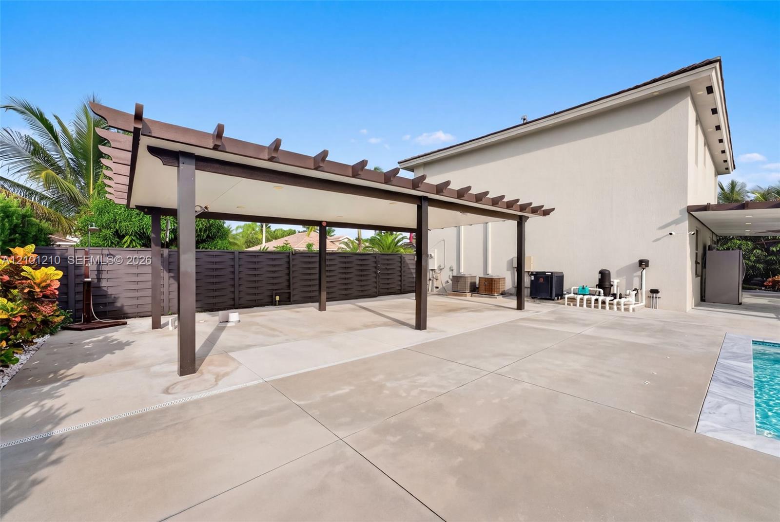 Modern home patio with a dark wood pergola, privacy fence, tropical plants, and swimming pool.