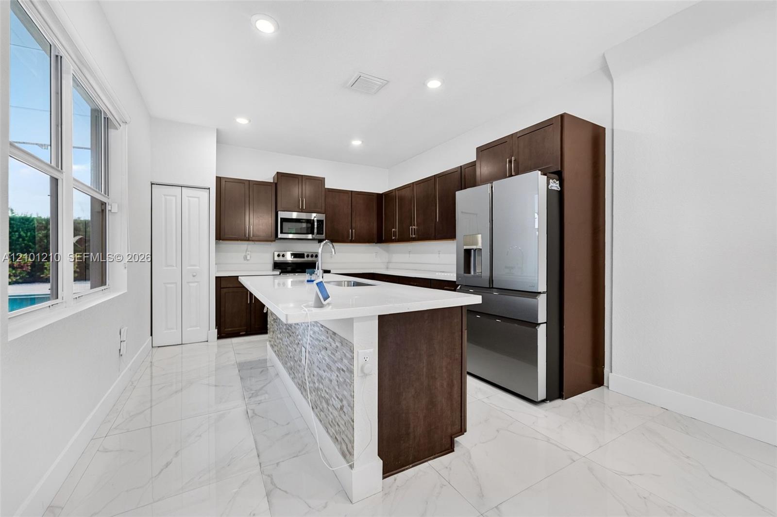 Kitchen with dark wood cabinets, a white island, stainless steel appliances, and large windows.