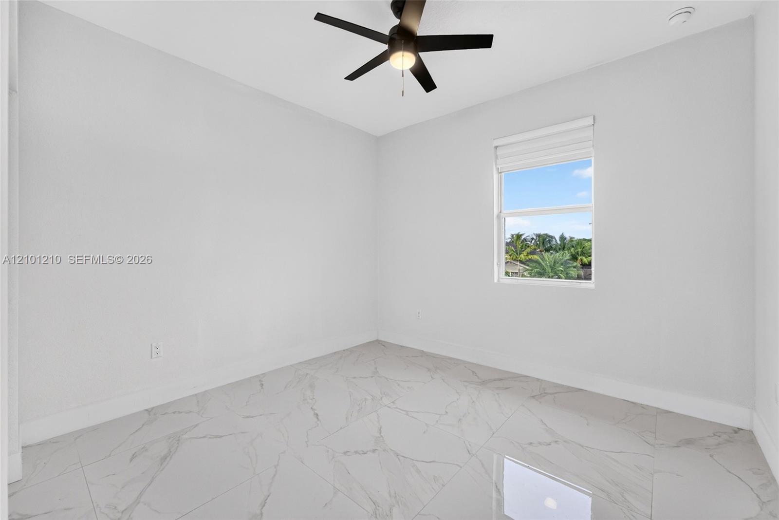 Empty white room with glossy marble-look tile floors, a black ceiling fan, and a window overlooking palms.