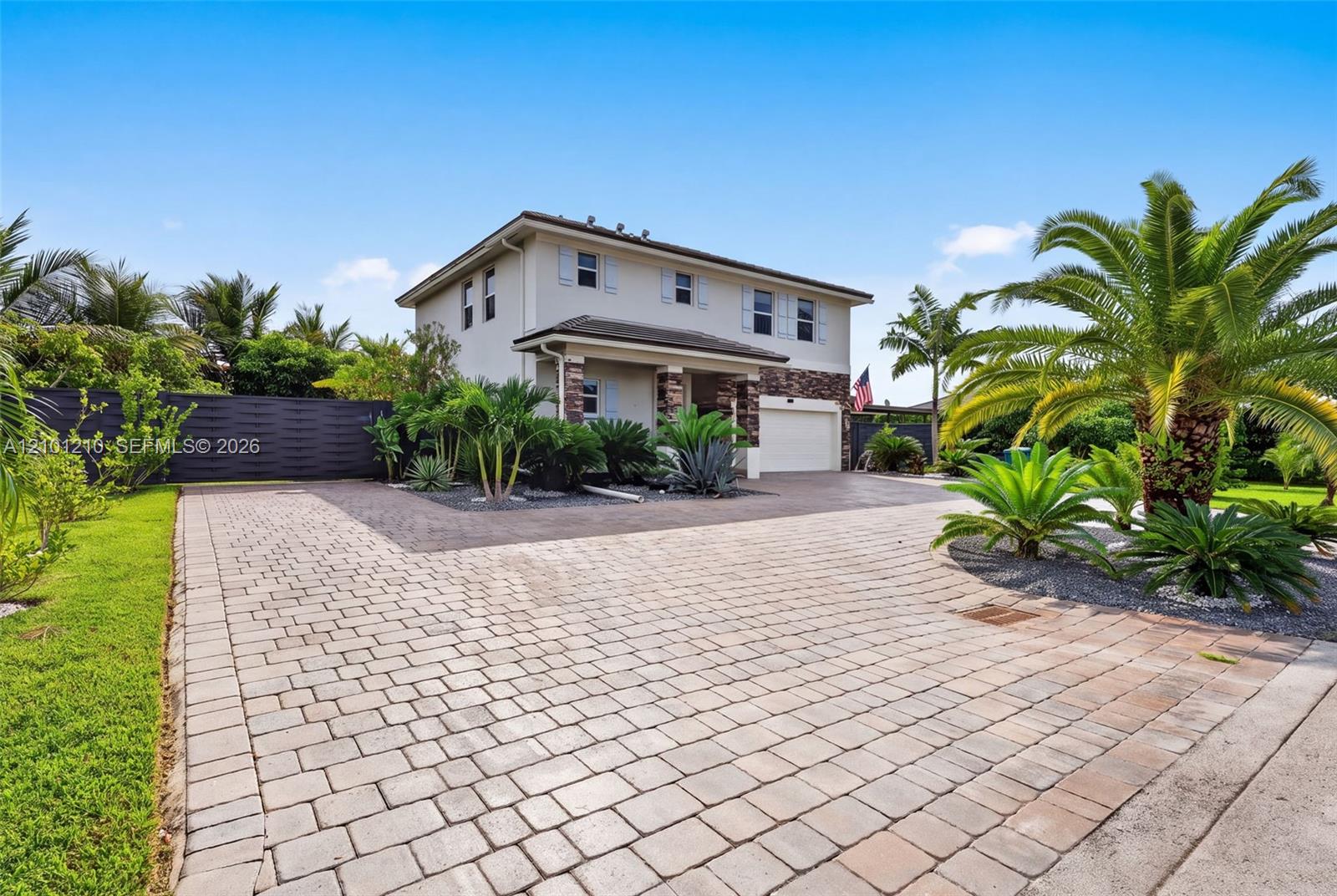 Two-story house with stone accents, a broad paver driveway, and tropical landscaping.