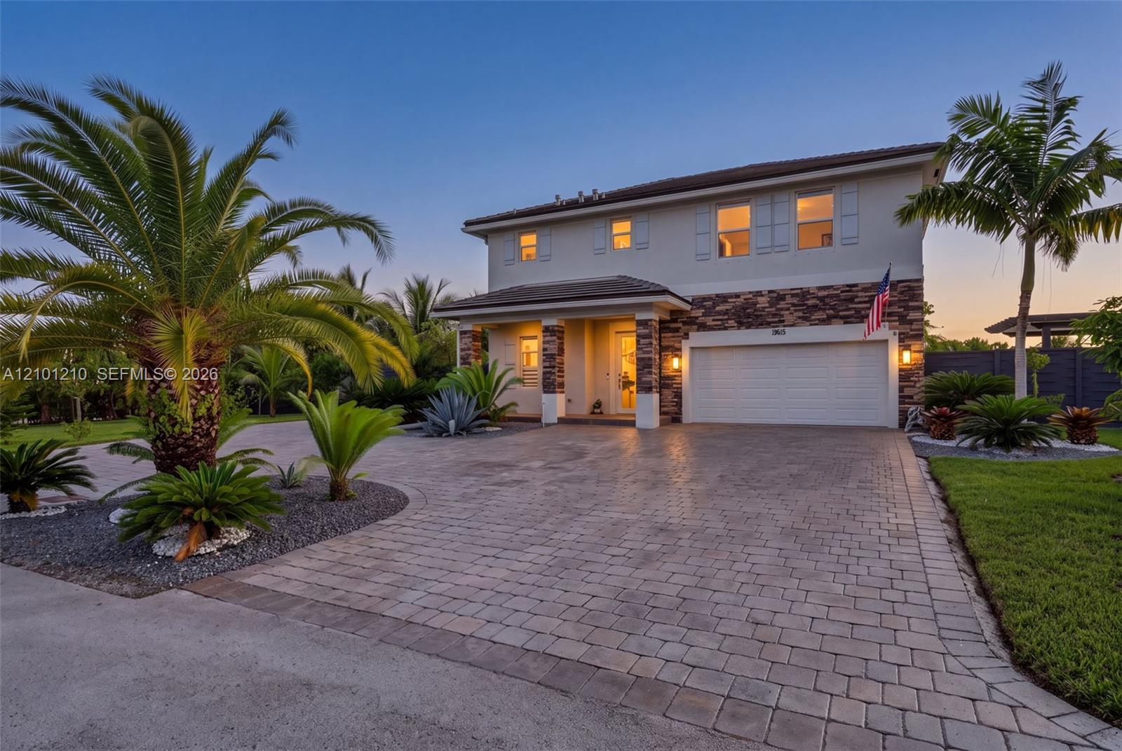 Two-story house with stone accents, a broad paver driveway, tropical landscaping, and warmly lit windows at dusk.