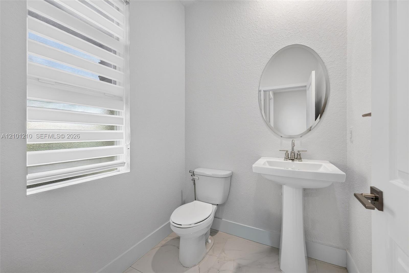 Bathroom with a pedestal sink, oval mirror, and striped window blinds beside the toilet.
