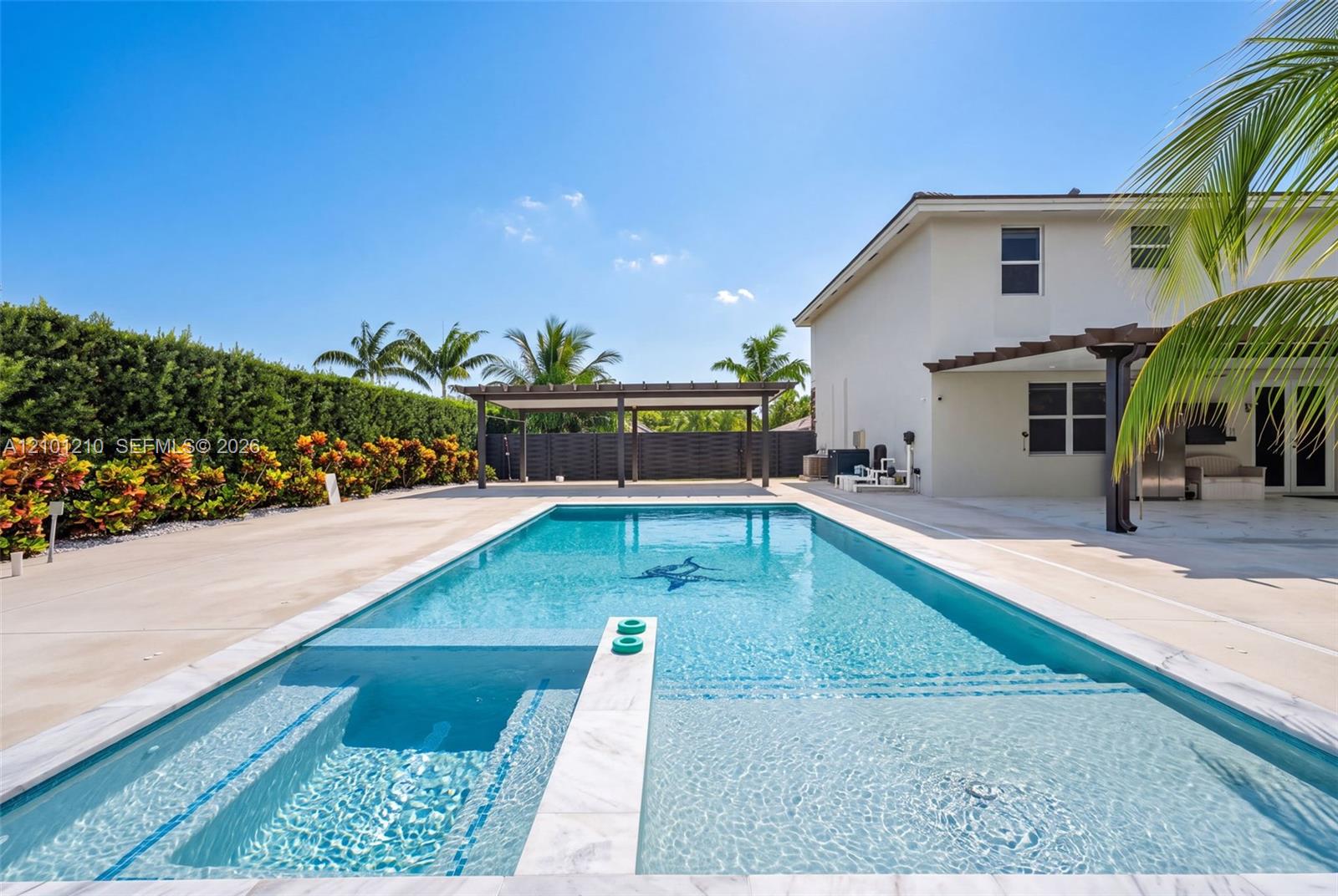 Backyard pool with a shallow tanning shelf, paved patio, pergola, and tropical landscaping beside a modern home.