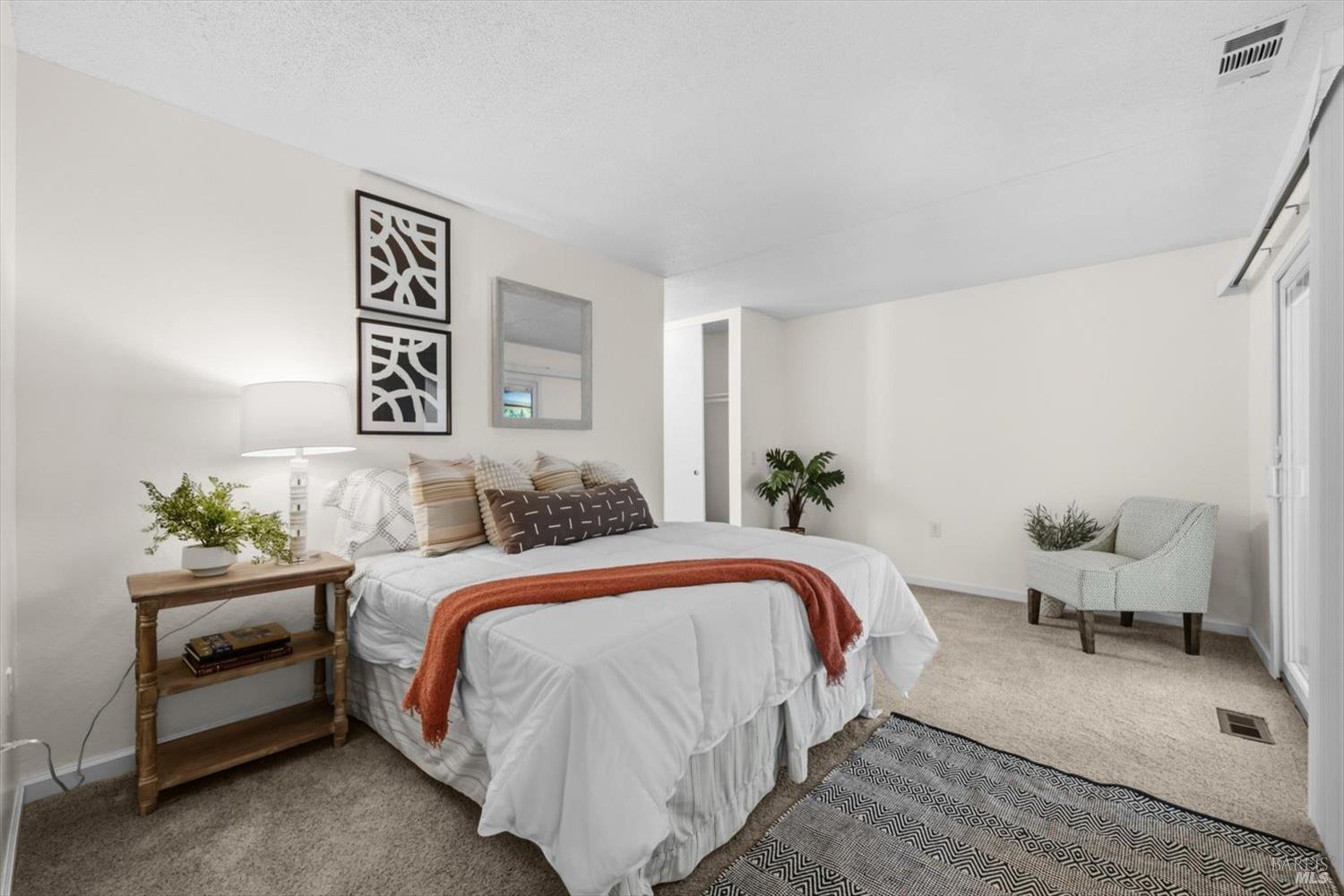 Staged bedroom with a white bed, rust-colored throw, wooden bedside table, and gray accent chair.