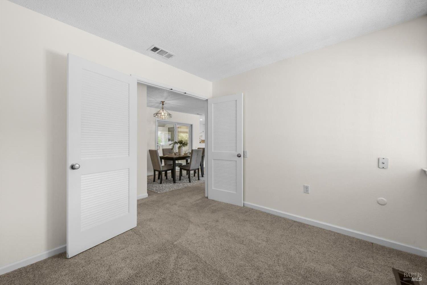 Carpeted room with open louvered doors looking into a furnished dining area.