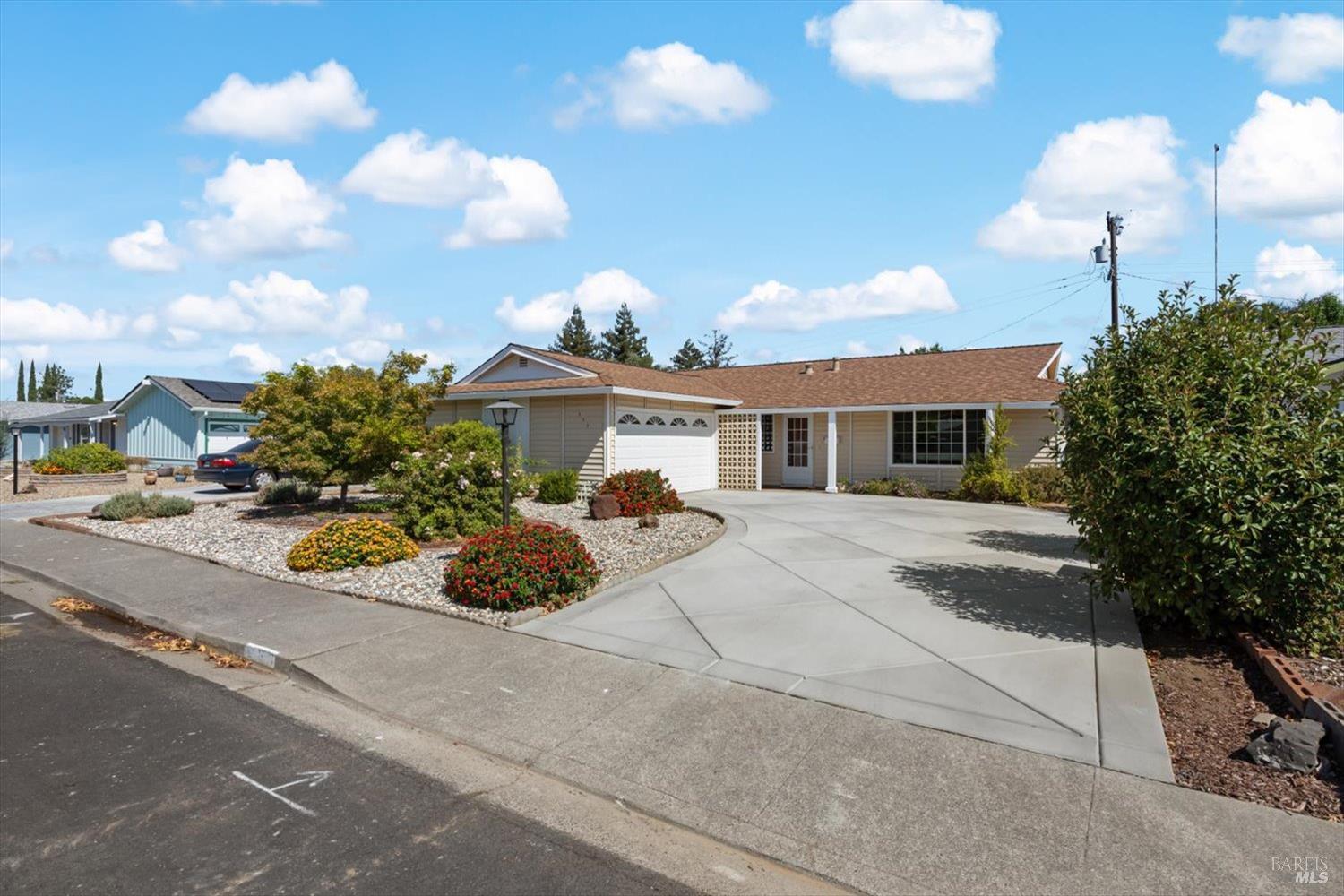 Single-story tan house with an attached garage, broad driveway, and flower-filled front garden.