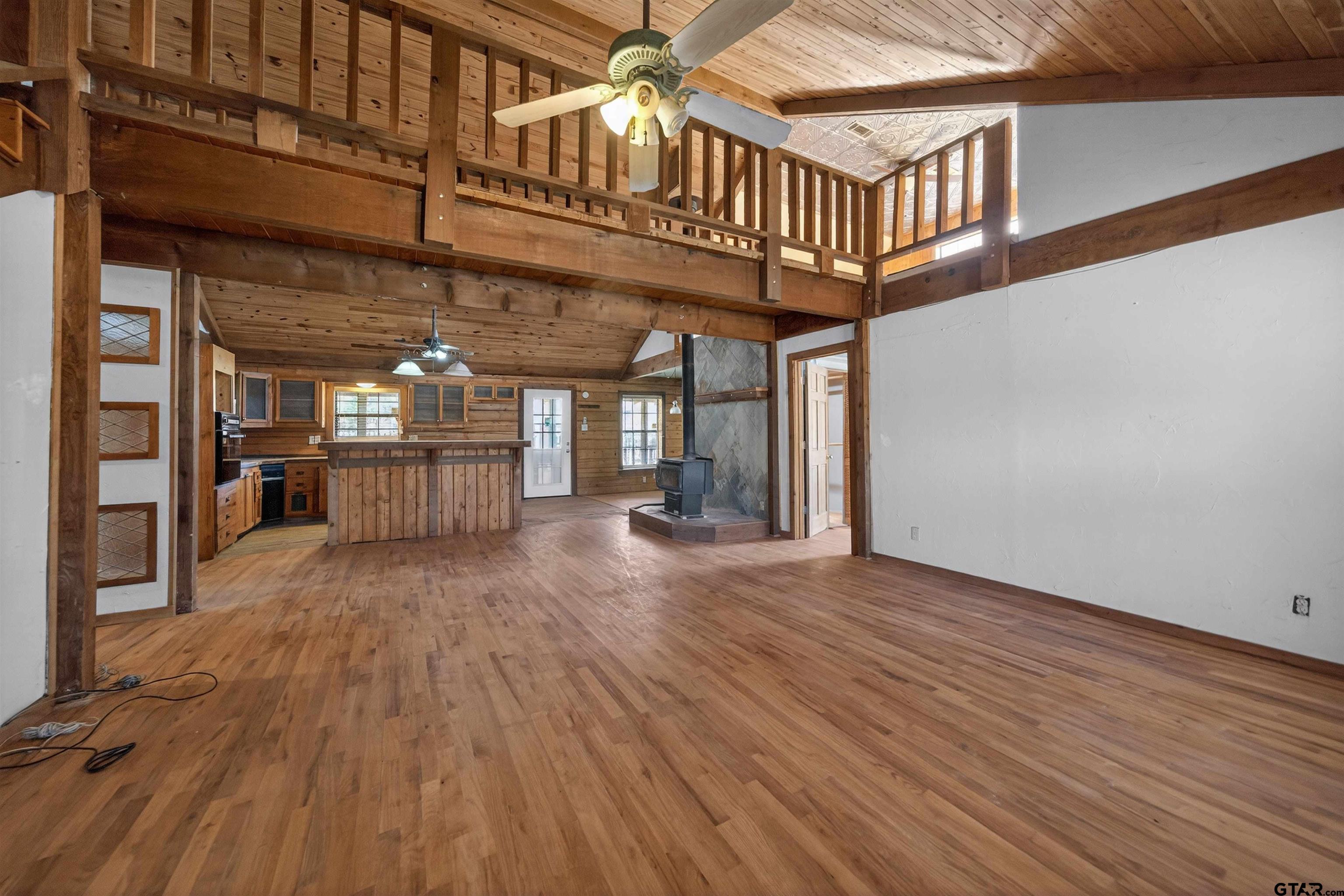 Open-plan living room with hardwood floors, wood-paneled loft, and freestanding stove.