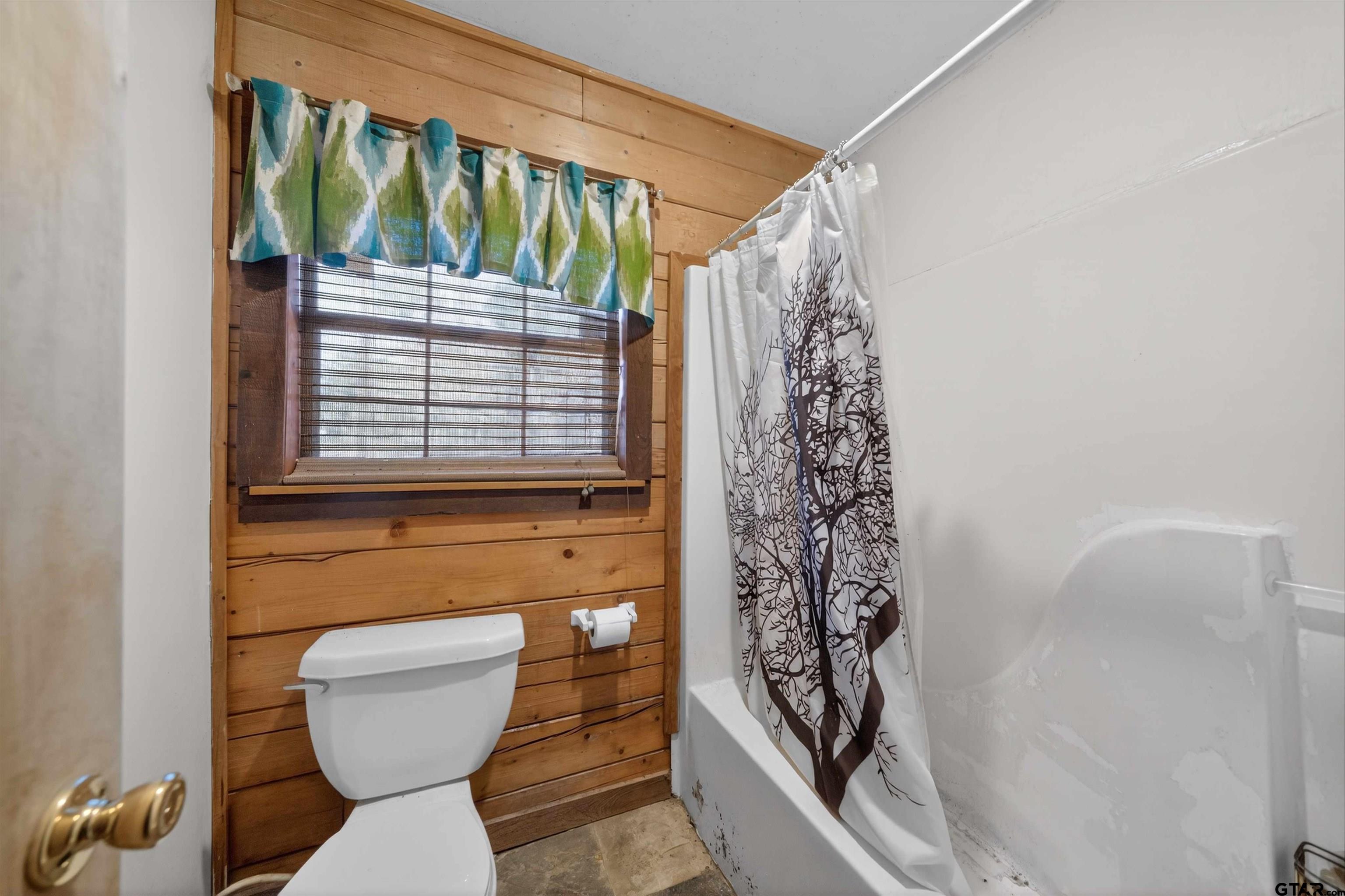 Bathroom with a white tub, tree-print shower curtain, and wood-paneled wall around the window.