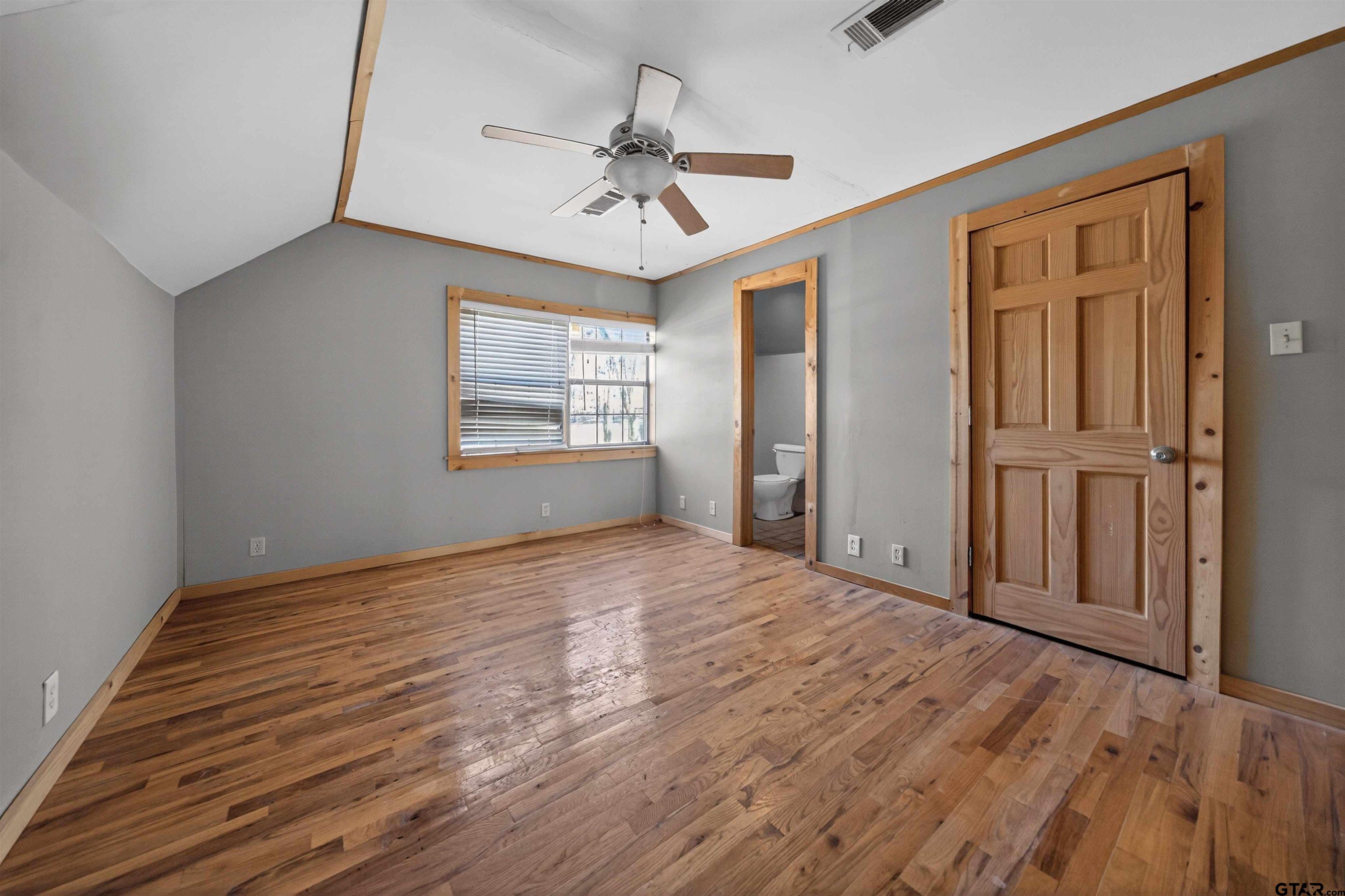 Vacant bedroom with hardwood floors, a vaulted ceiling, and a bathroom visible through the doorway.