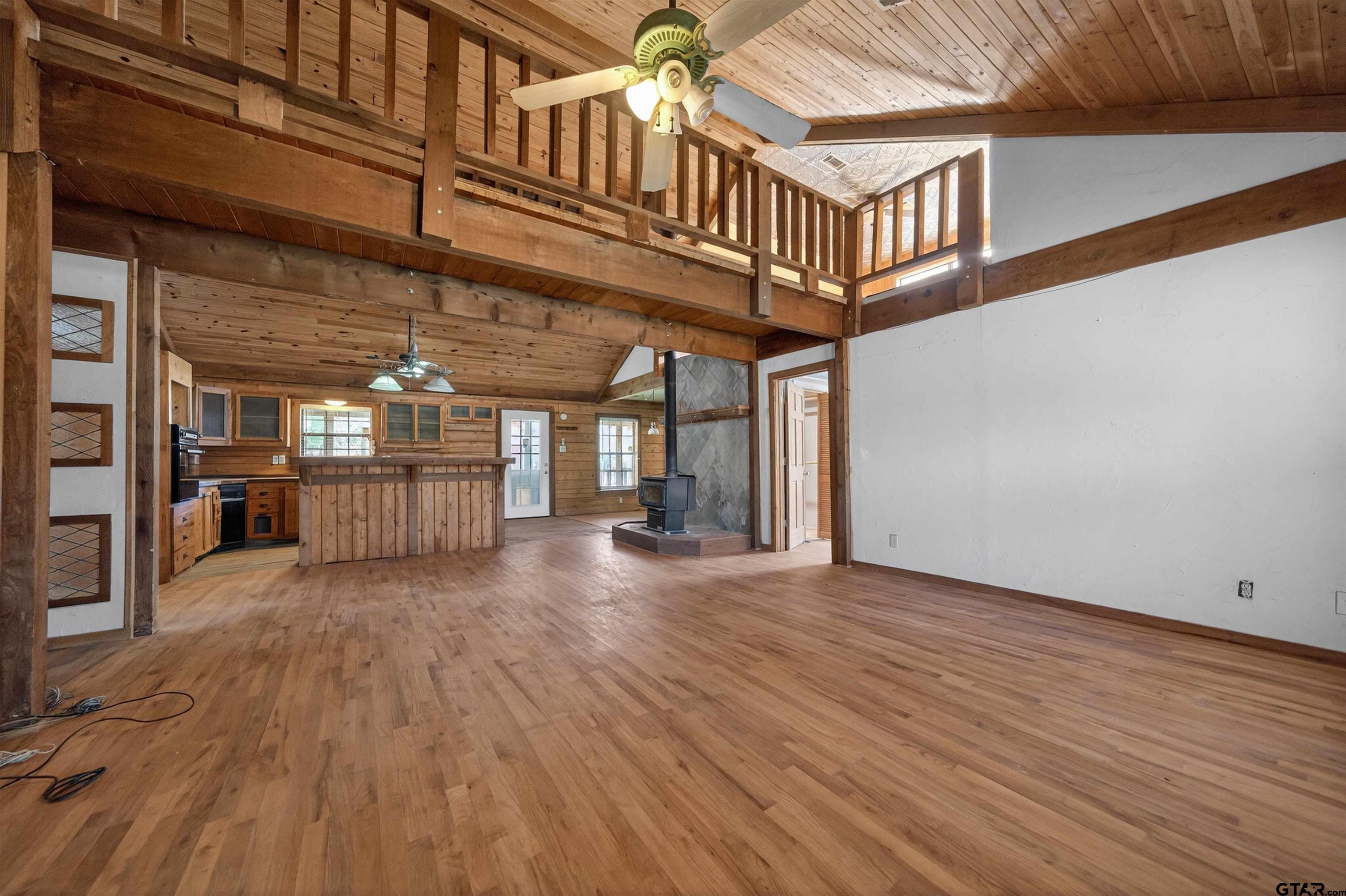 Open living room with a wood-railed loft, wood-plank ceiling, kitchen island, and freestanding stove.