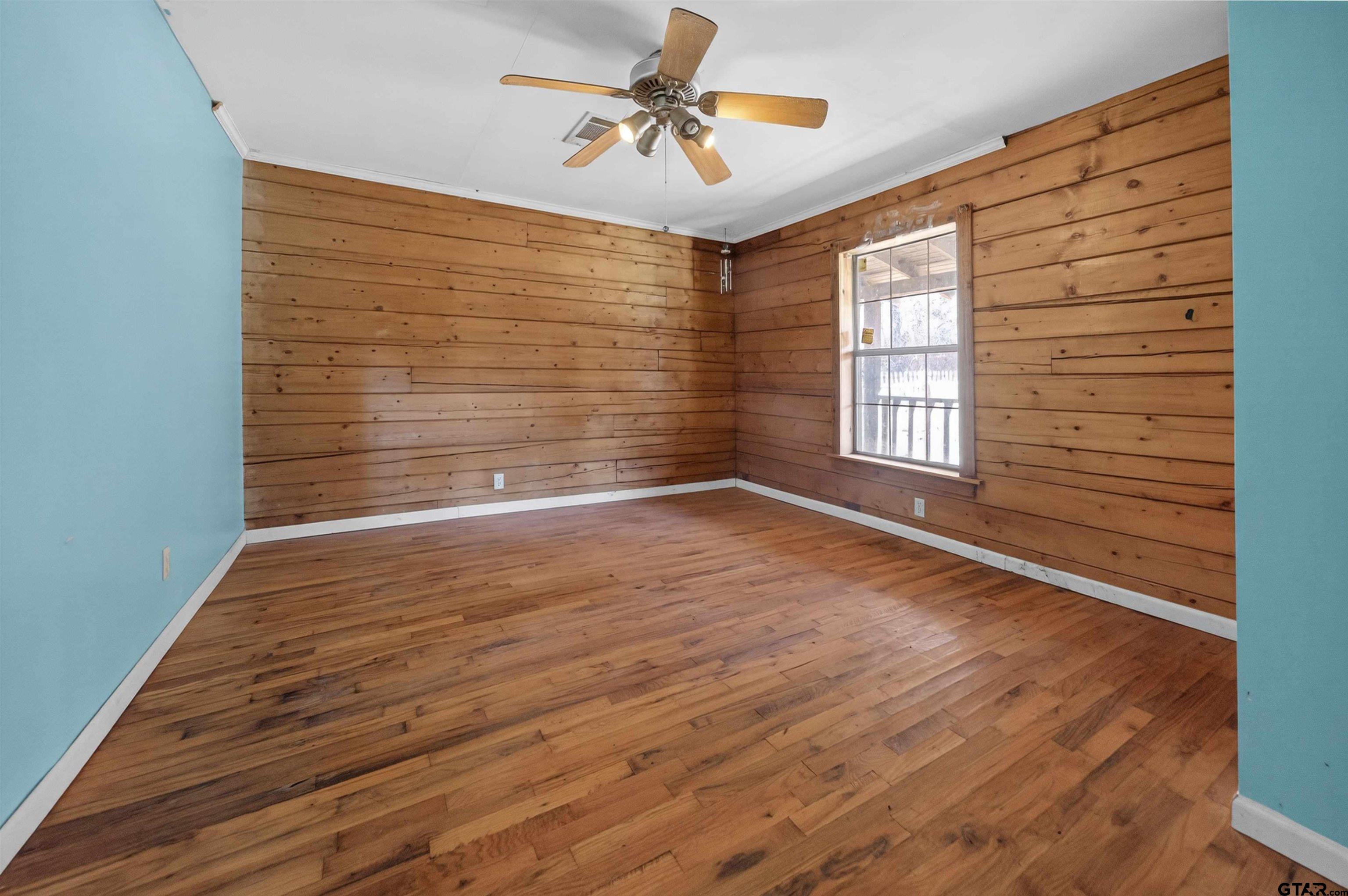 Empty room with hardwood floors, wood-paneled walls, a ceiling fan, and a window.