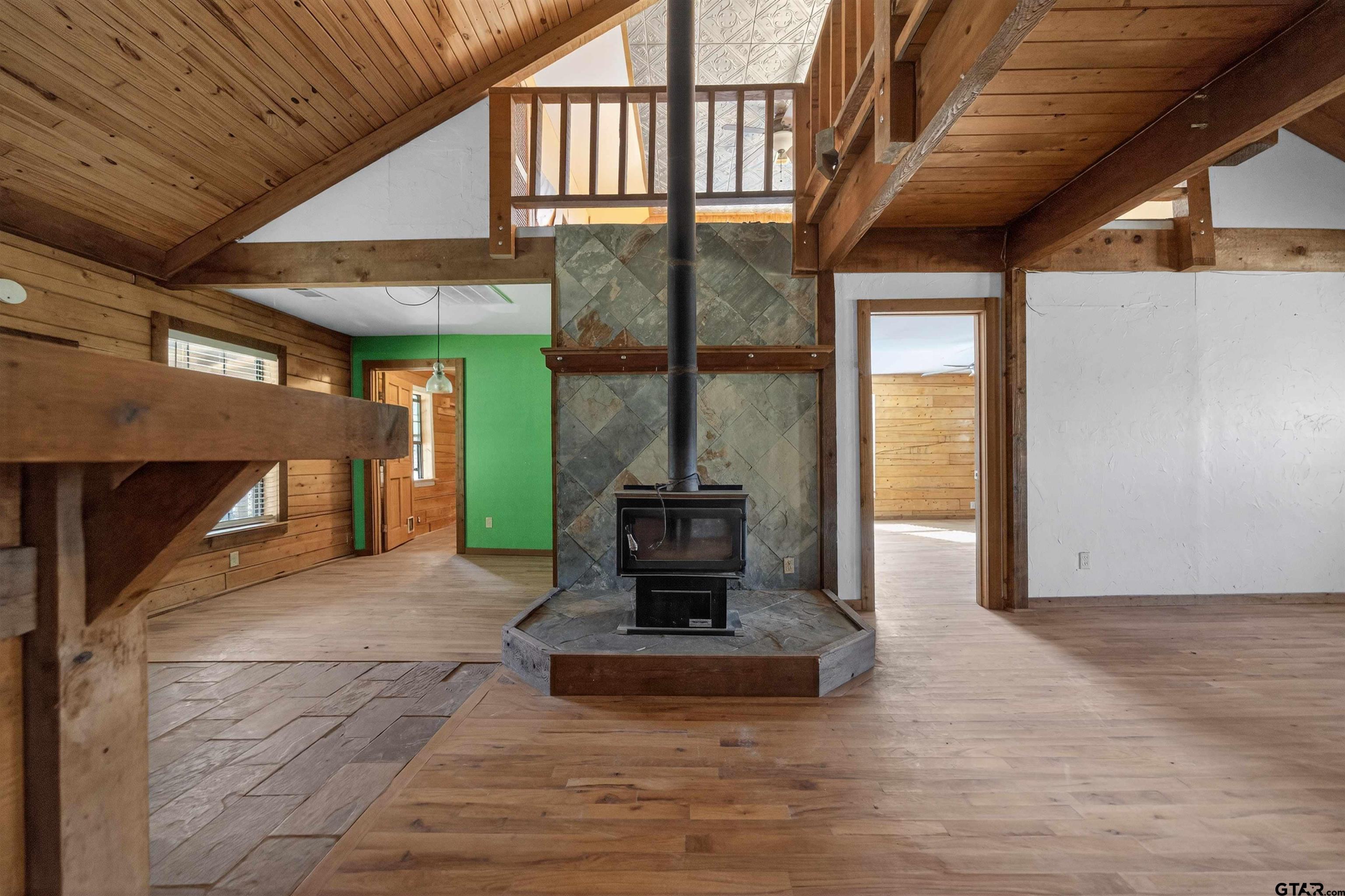 Vacant living room with vaulted wood ceilings, exposed beams, and a freestanding stove with stone surround.