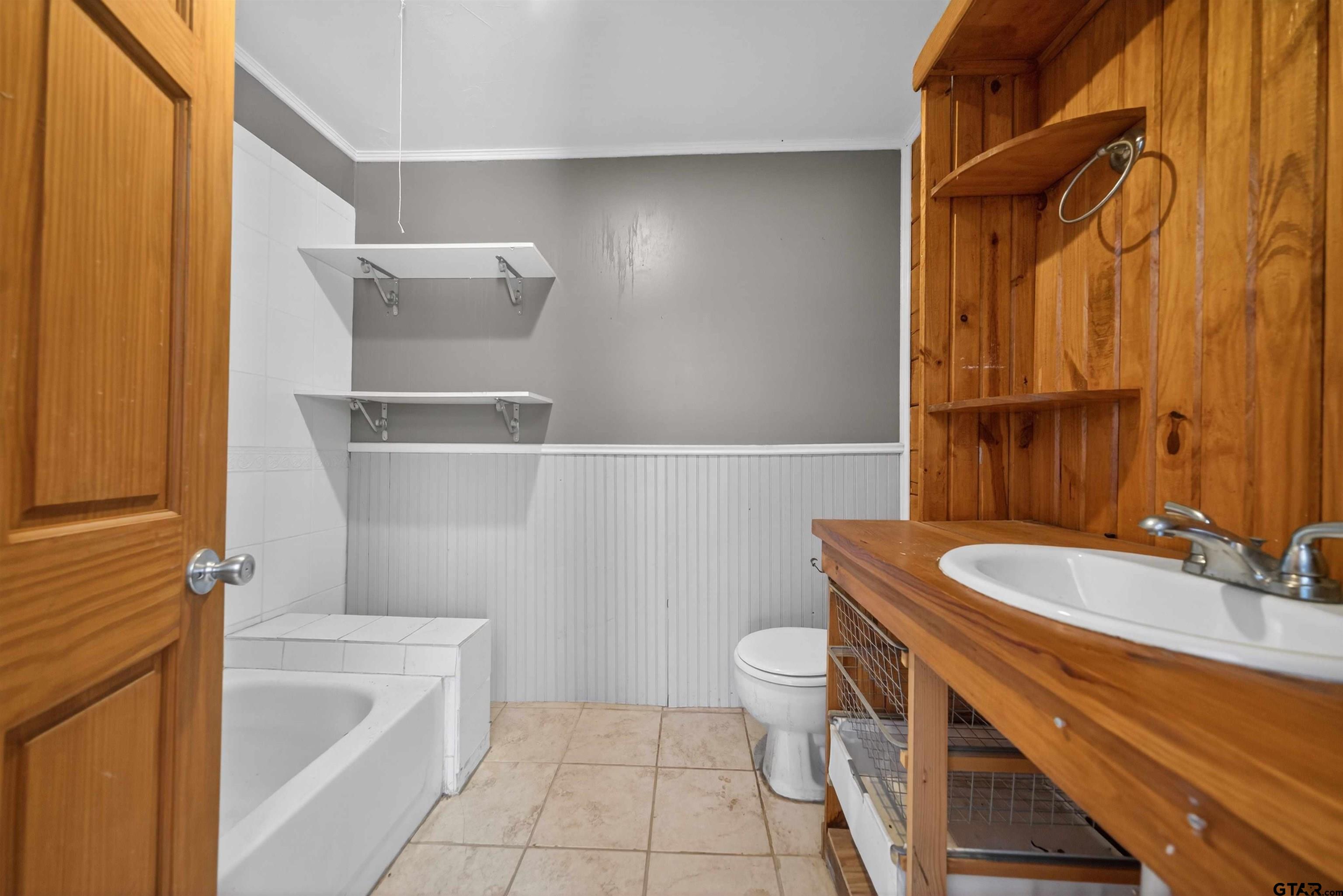 Bathroom with a white tub and toilet, beadboard wainscoting, and a wooden vanity with open shelving.