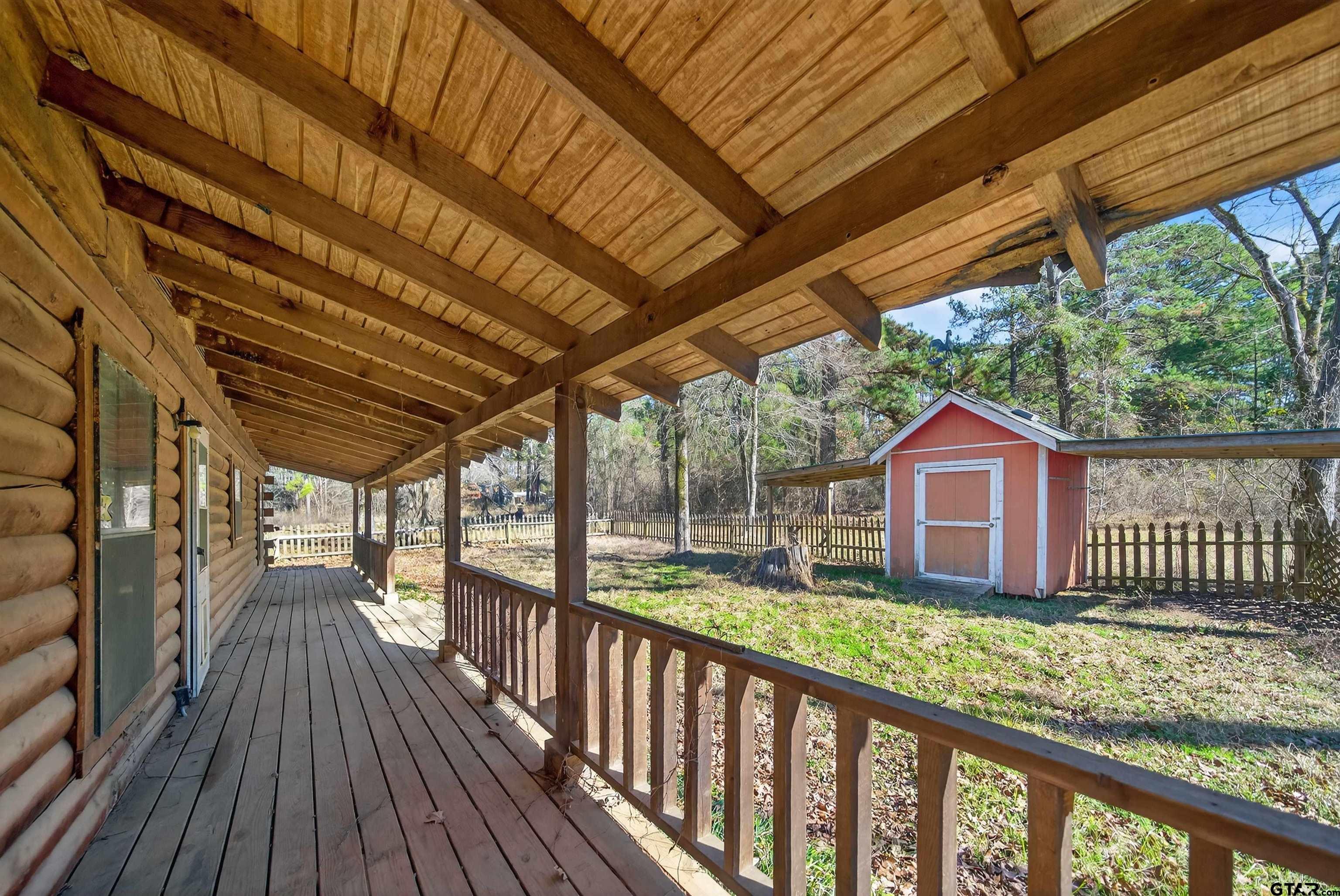 Covered timber porch beside a log home, overlooking a fenced yard and red shed.
